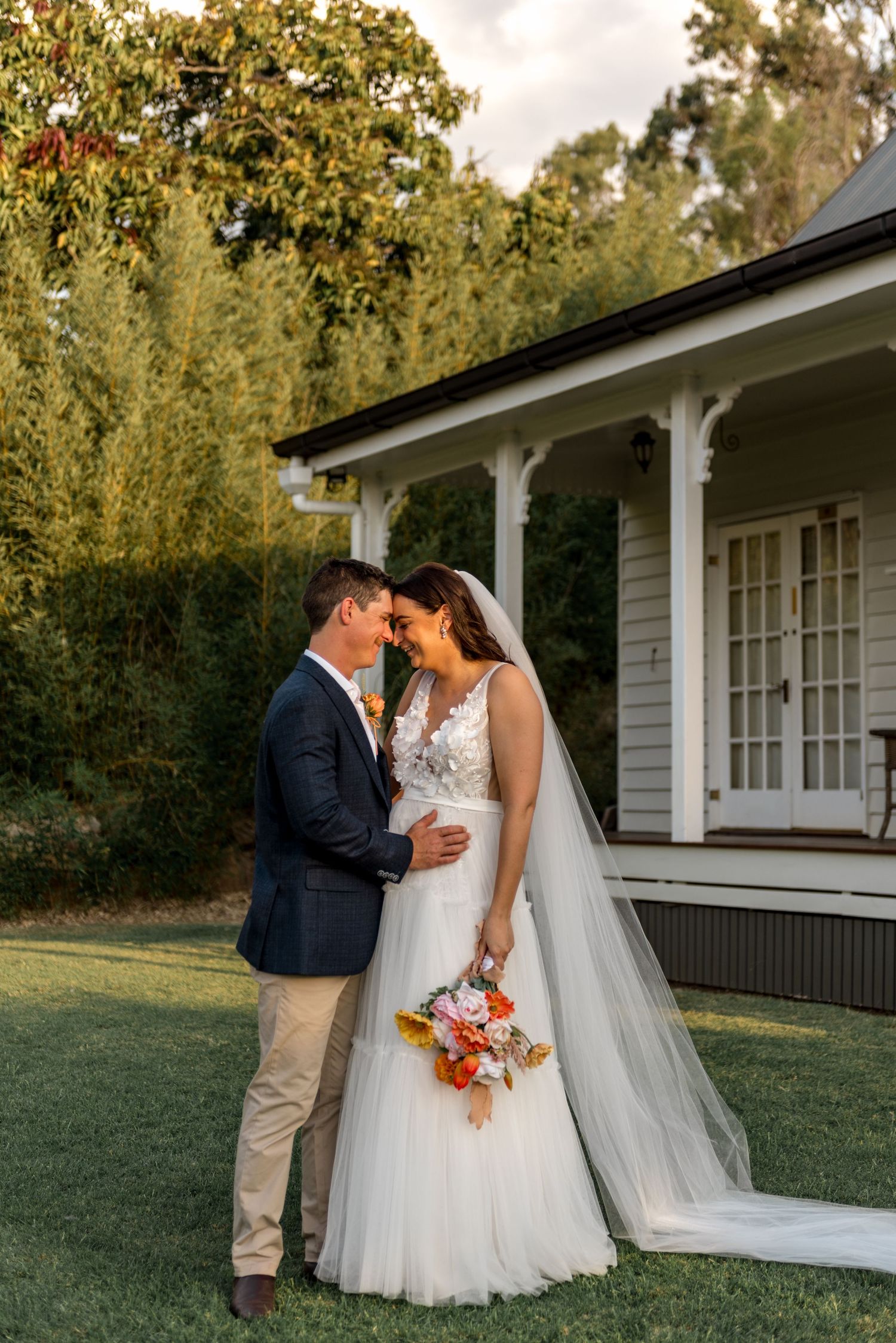 Bride and groom touching foreheads tenderly outside a white country farmhouse at golden hour.