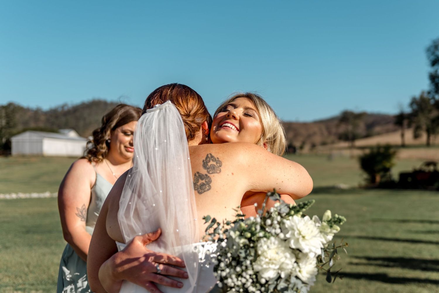 Bride hugging smiling bridesmaid in sage green dress outdoors on green lawn with blue sky.