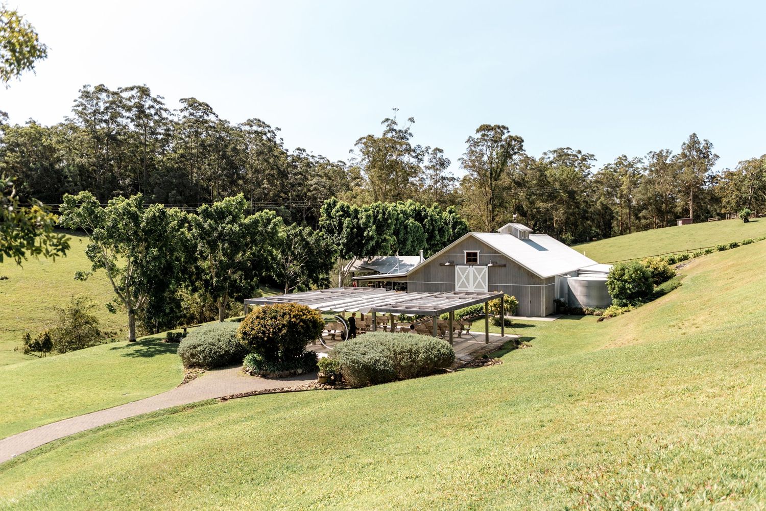 Rustic wedding venue with white barn, pergola, and lush green hillside surrounded by tall trees on a sunny day.
