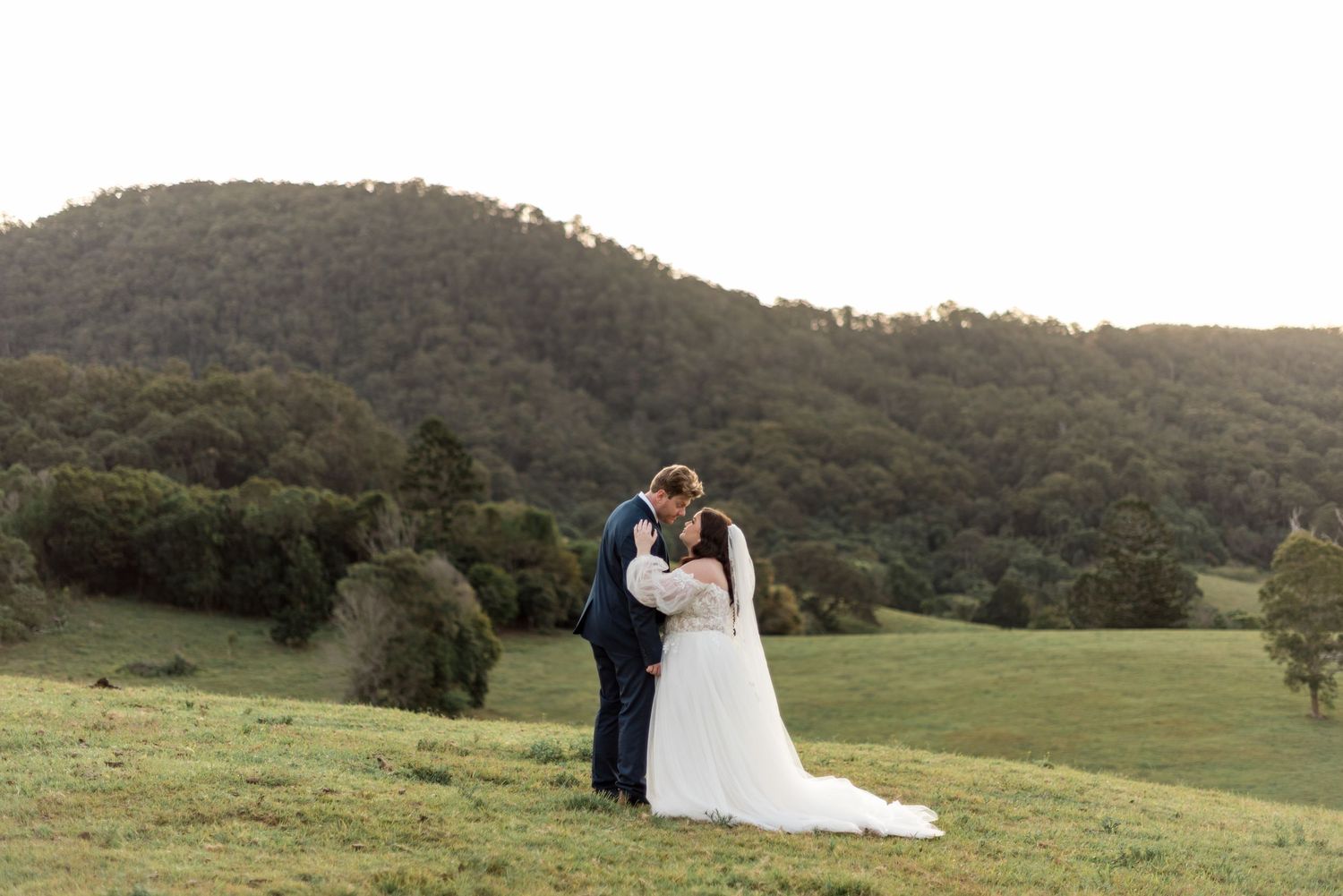 Bride and groom share a romantic kiss on a lush green hillside with rolling mountains in the background.