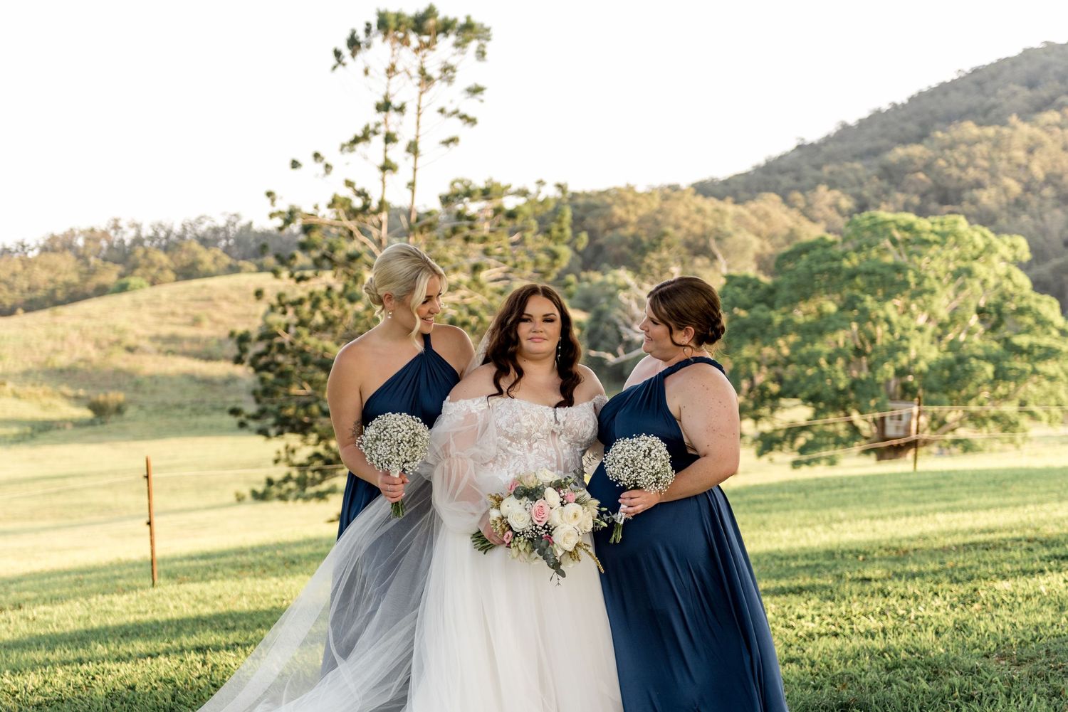 Bride with two bridesmaids in navy dresses holding bouquets on green hillside at golden hour.