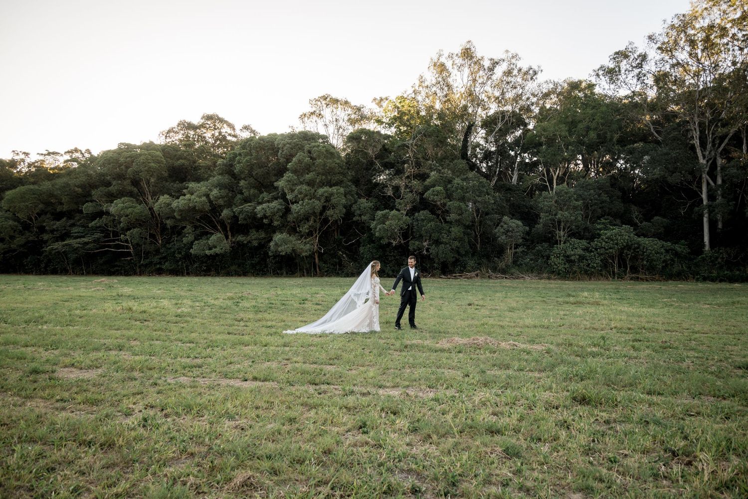 Bride and groom walk hand in hand across an open green field with tall eucalyptus forest backdrop.