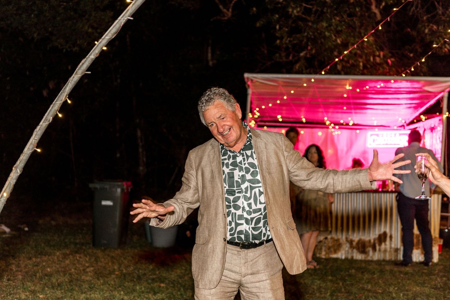 Cheerful older man in a patterned blazer dances energetically at an outdoor evening wedding reception.