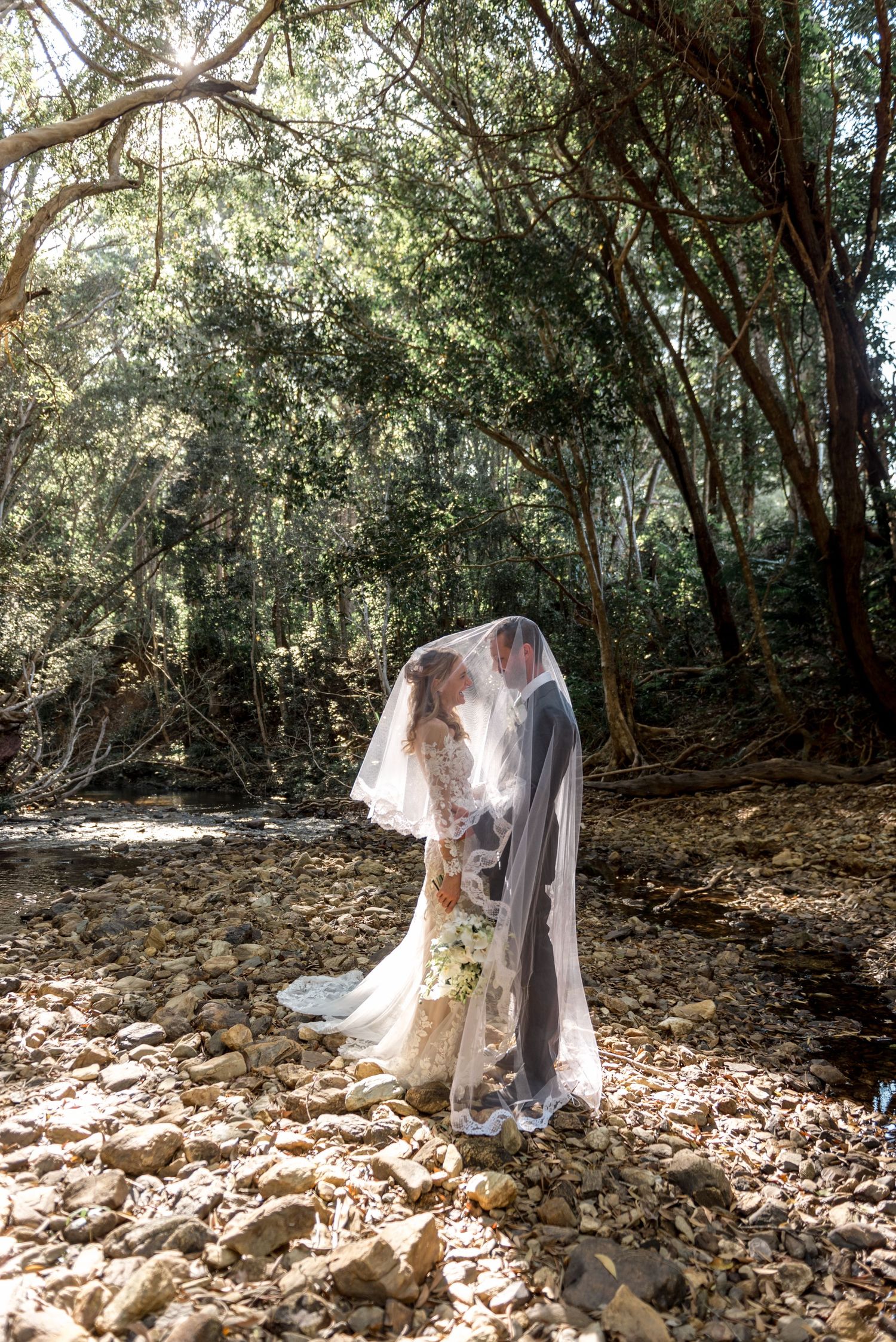 Bride and groom kissing under flowing veil on rocky creek bed in sunlit forest setting.