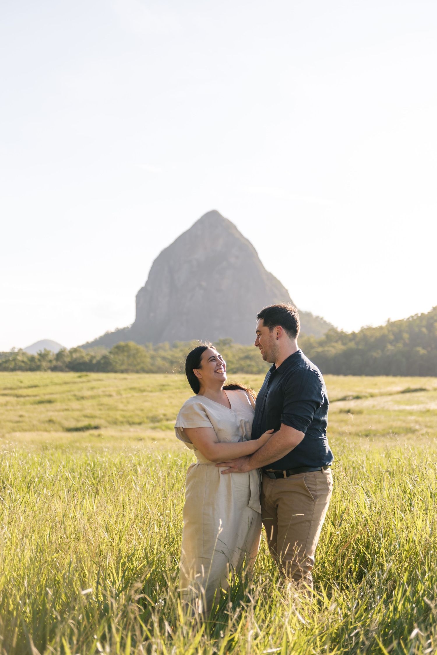 Happy couple holding each other in a sunlit green field with a large rocky mountain in the background.