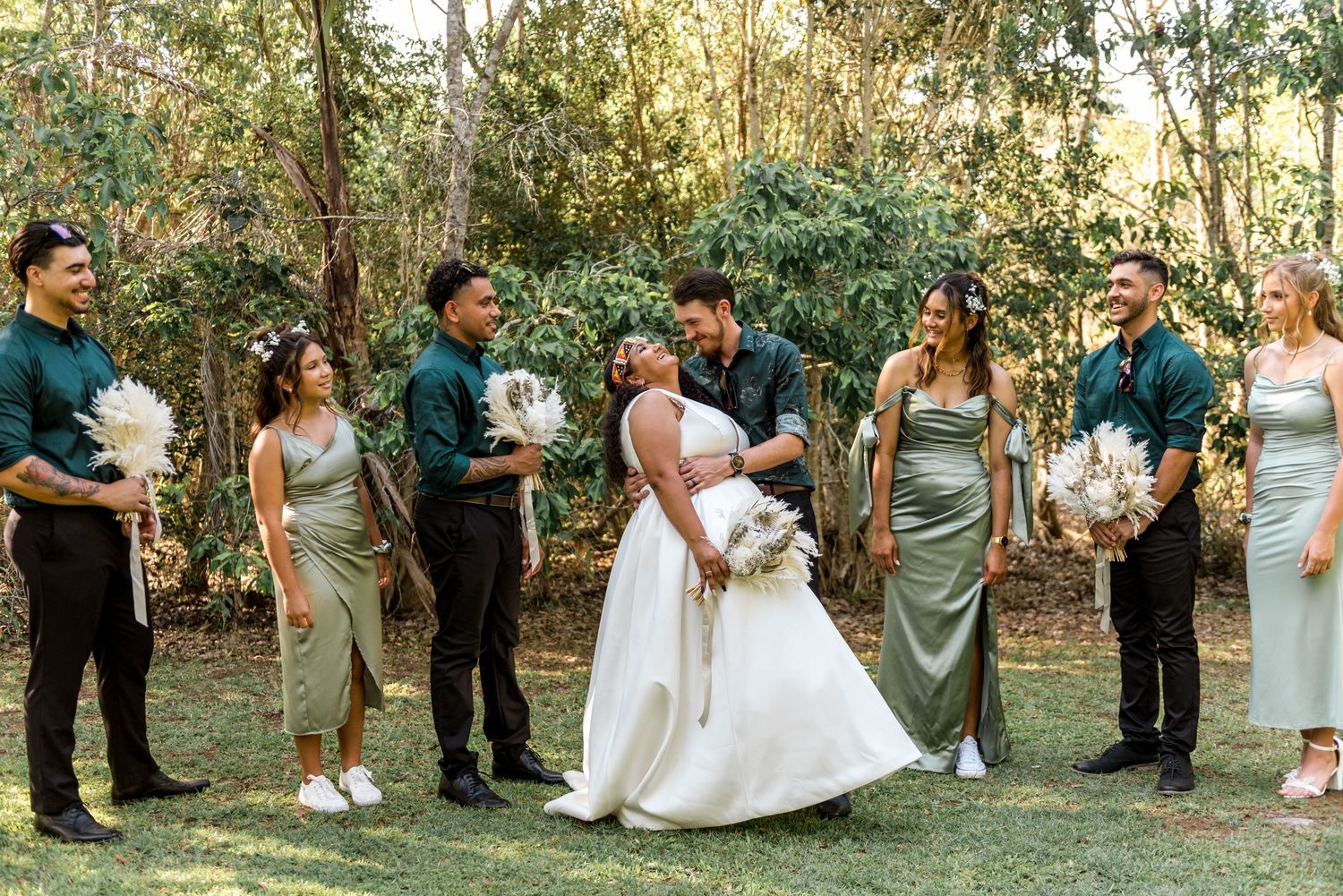 A bride and groom kiss while their wedding party in green attire surrounds them outdoors.