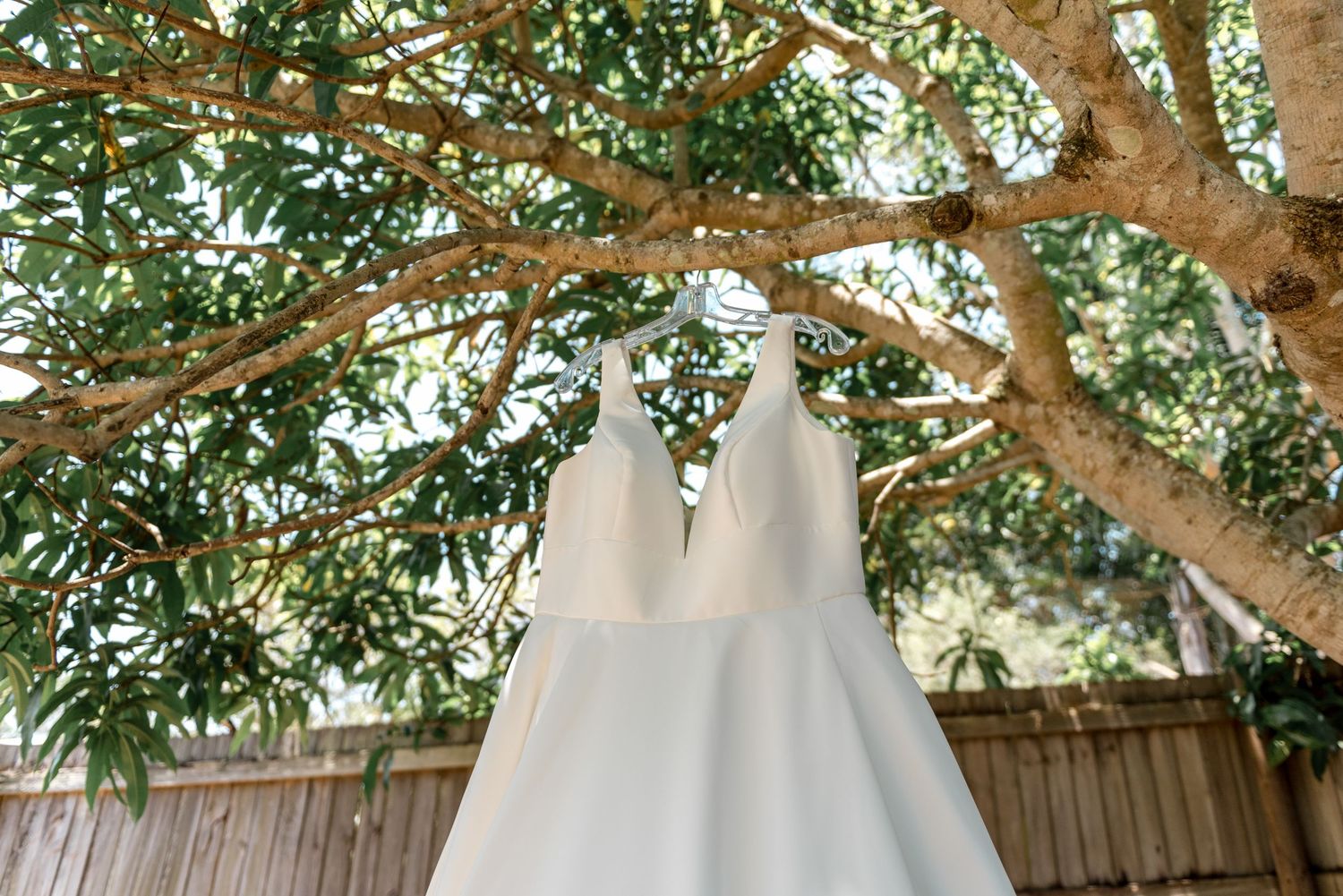 Elegant white wedding dress hanging from a tree branch outdoors against a wooden fence backdrop.