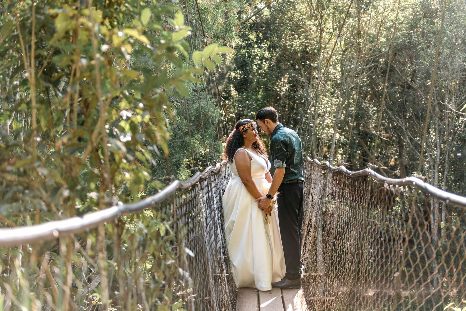 A bride and groom share an intimate moment on a rope bridge surrounded by green foliage.
