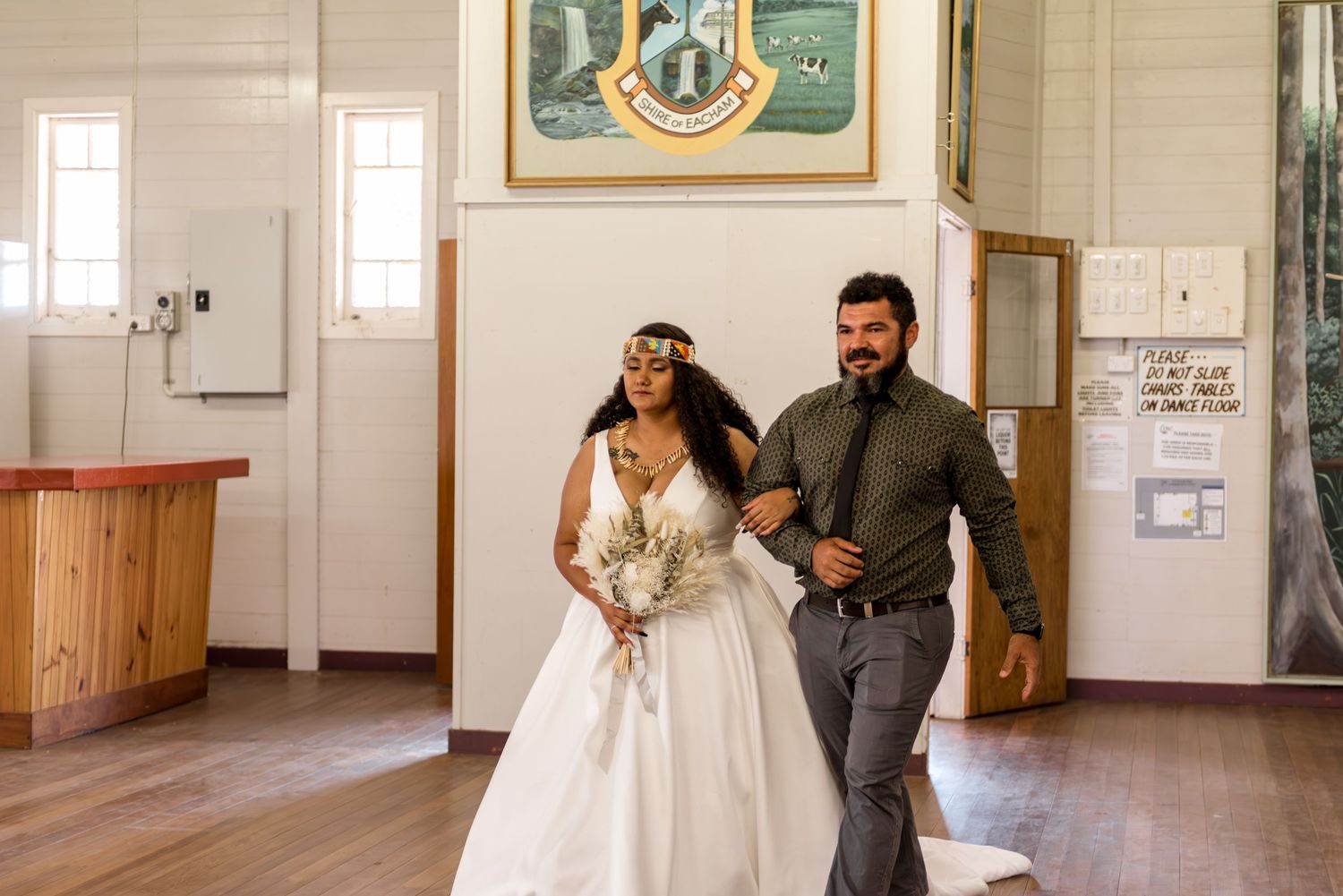 Bride in white ball gown with colorful headpiece walks arm-in-arm with groomsman into rustic wedding venue.