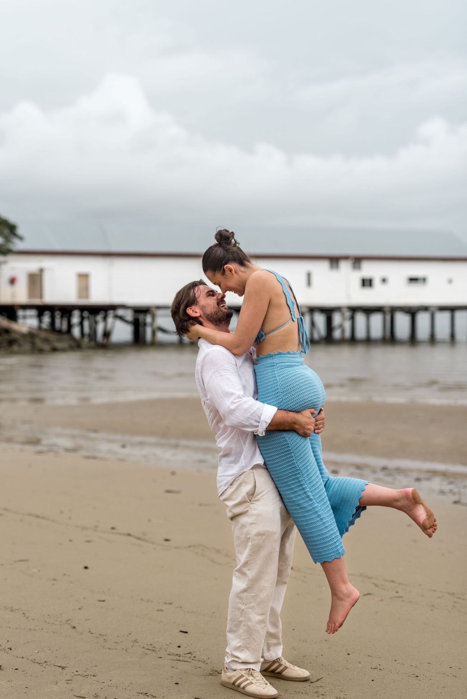 Man lifting woman for a kiss on a sandy beach with a pier in the background on a cloudy day.