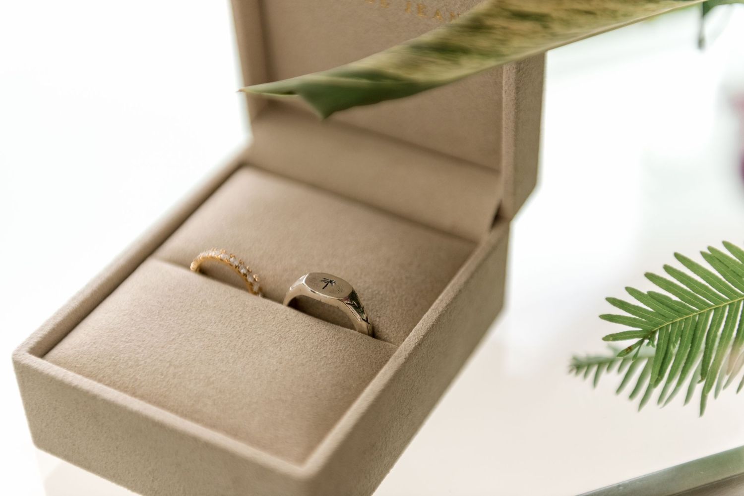 Two gold wedding bands displayed in a beige suede ring box with tropical greenery accent.
