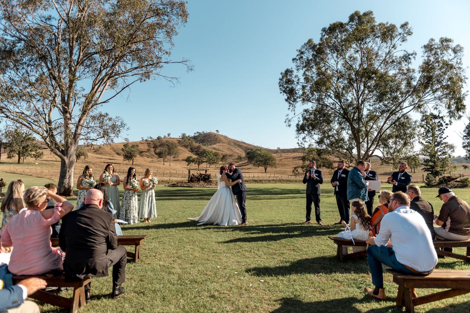 Wide shot of outdoor wedding ceremony on green lawn with guests seated on wooden benches and rolling hills behind.