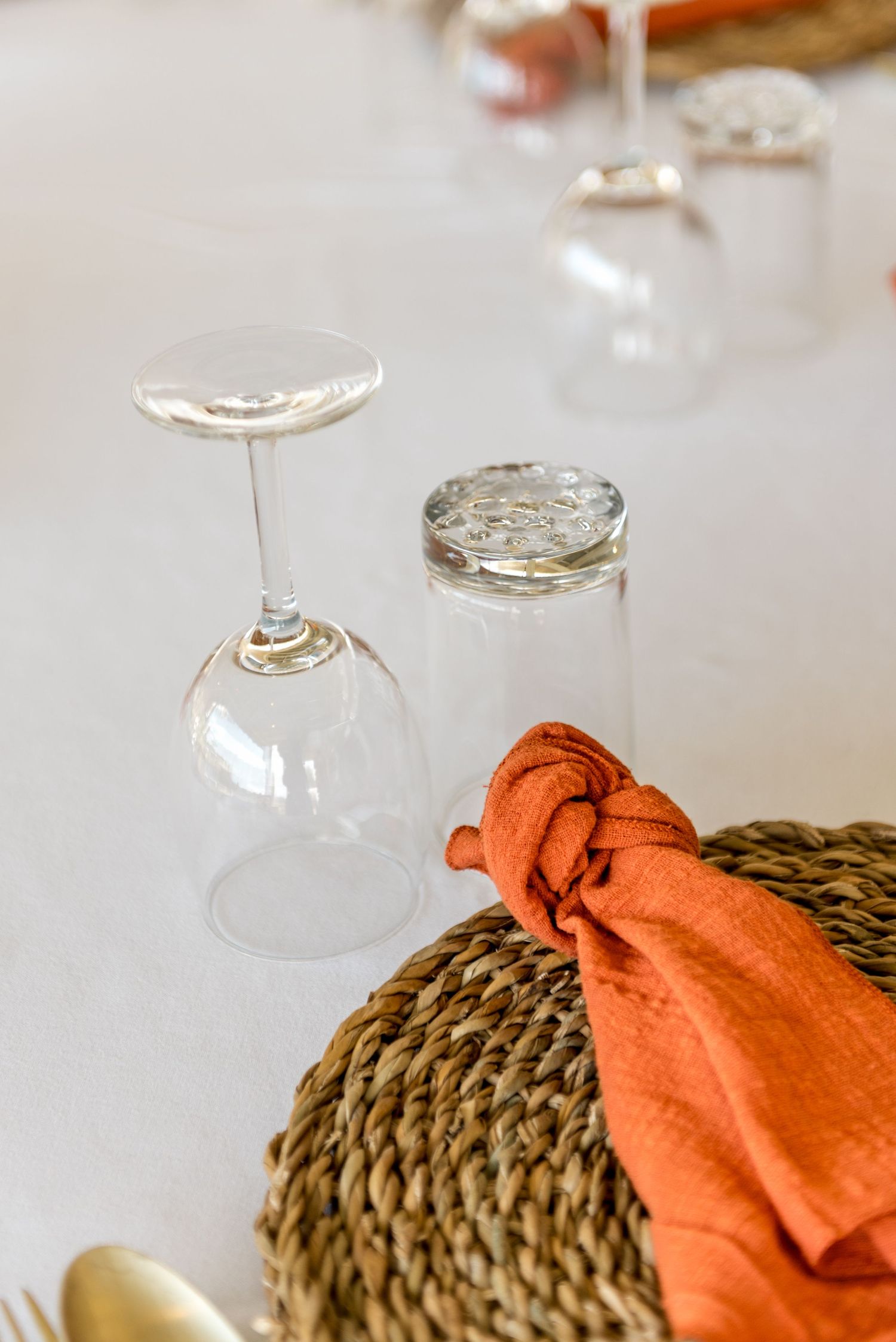 Wedding table setting with upturned wine glass, woven charger plate, and knotted burnt orange linen napkin on white tablecloth.