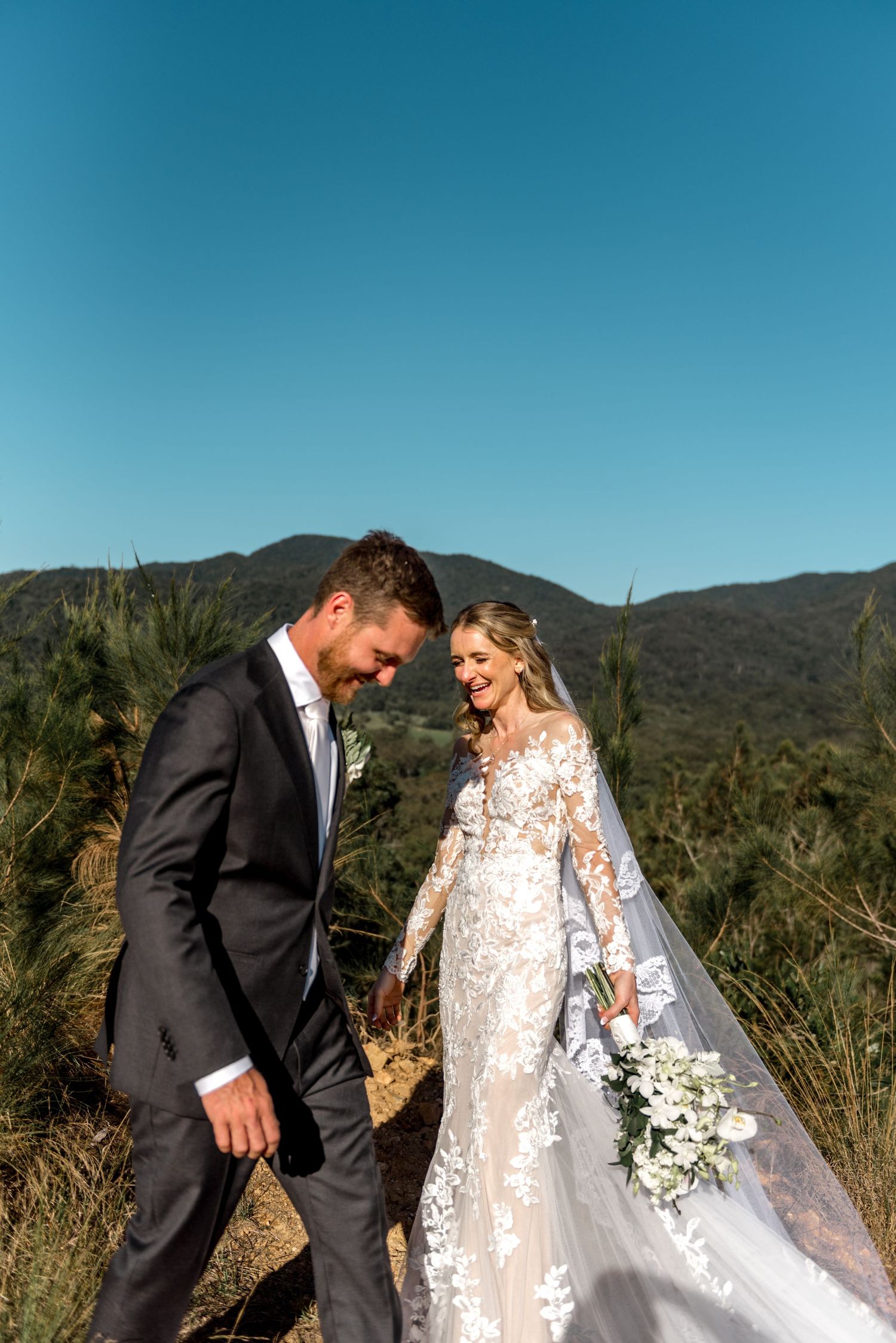 Bride and groom walking hand in hand through golden grass with mountain backdrop and blue sky.