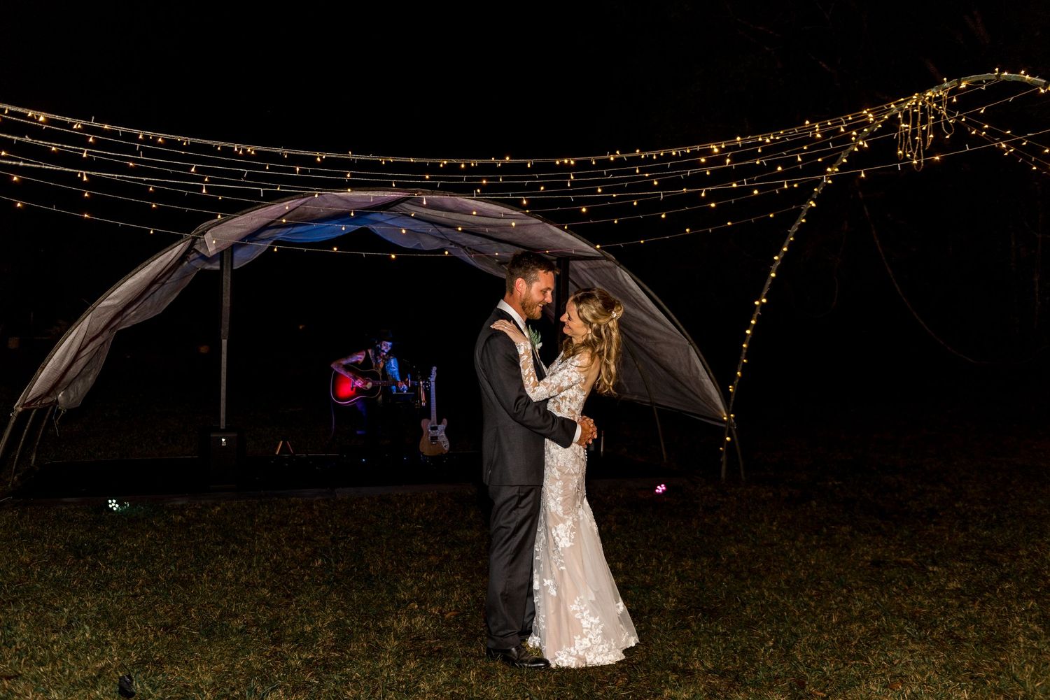 Bride and groom embrace under a glowing arch of fairy lights at their romantic outdoor nighttime wedding.