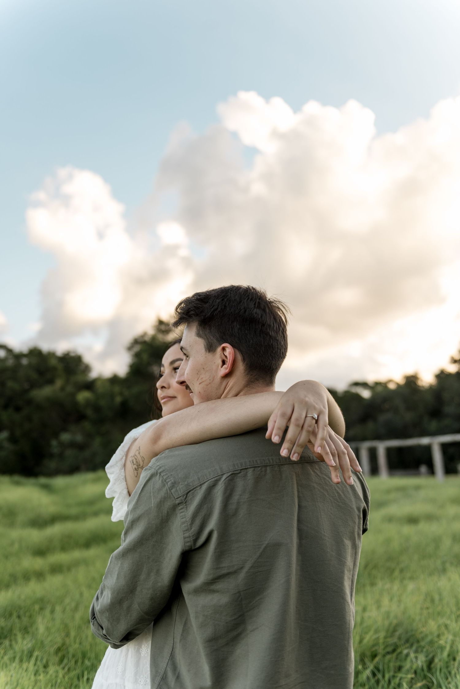 Couple embracing in green field at sunset, woman showing engagement ring while resting arms on mans shoulders.