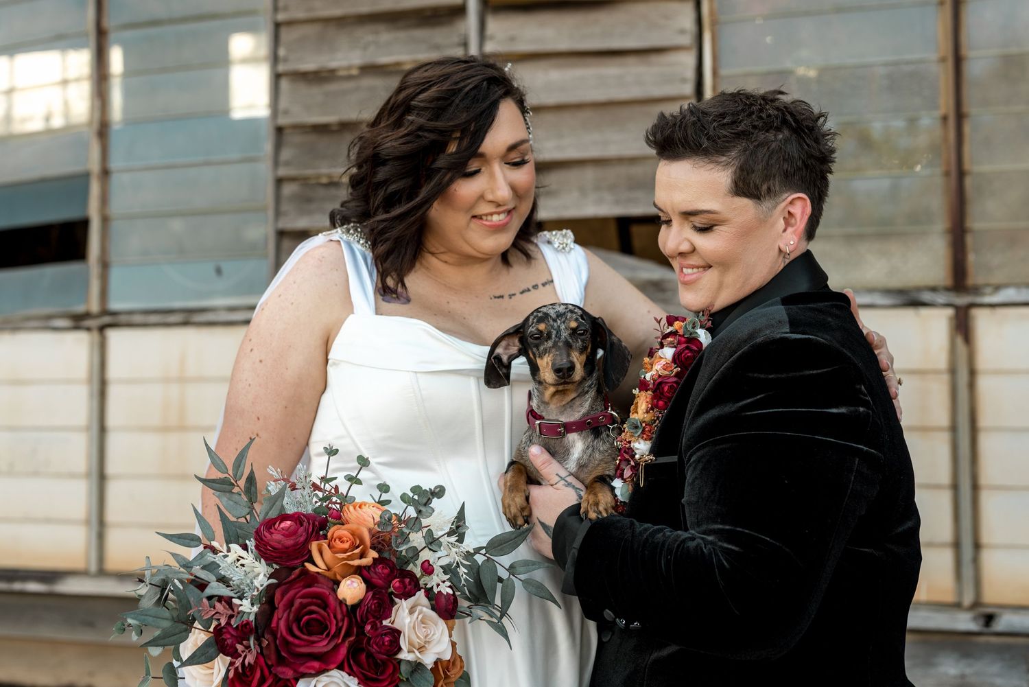 Two brides laugh together holding a cute dachshund dog with a boho floral bouquet at their wedding.
