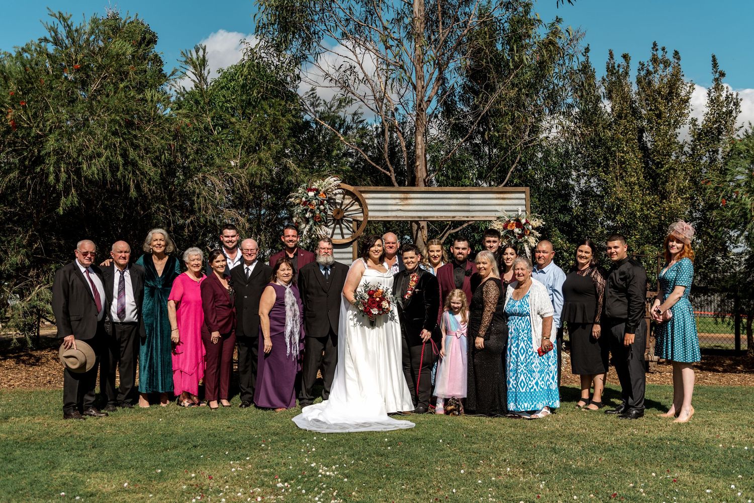 Wedding party group photo outdoors with bride and groom centered before a rustic floral arch on a sunny day.