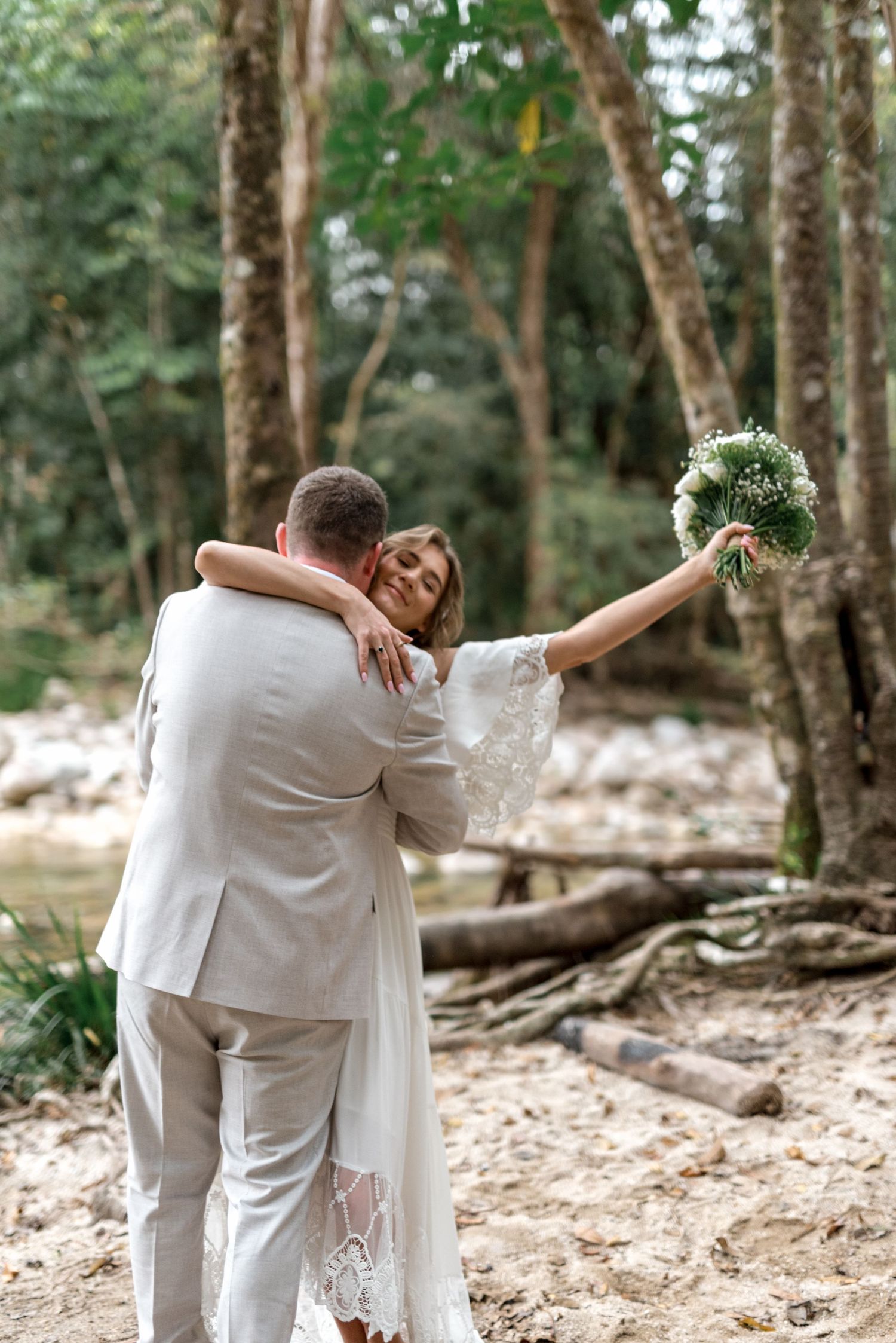 Joyful bride raises bouquet while embracing groom in a sun-dappled forest wedding celebration.
