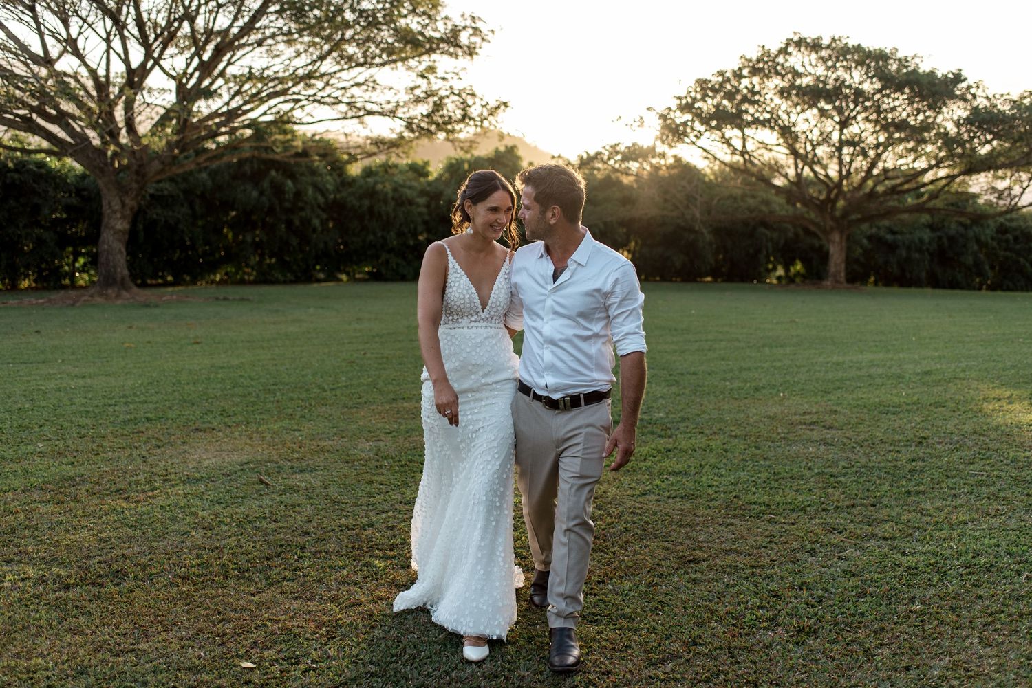 Smiling bride and groom walking together on green lawn during golden hour sunset.