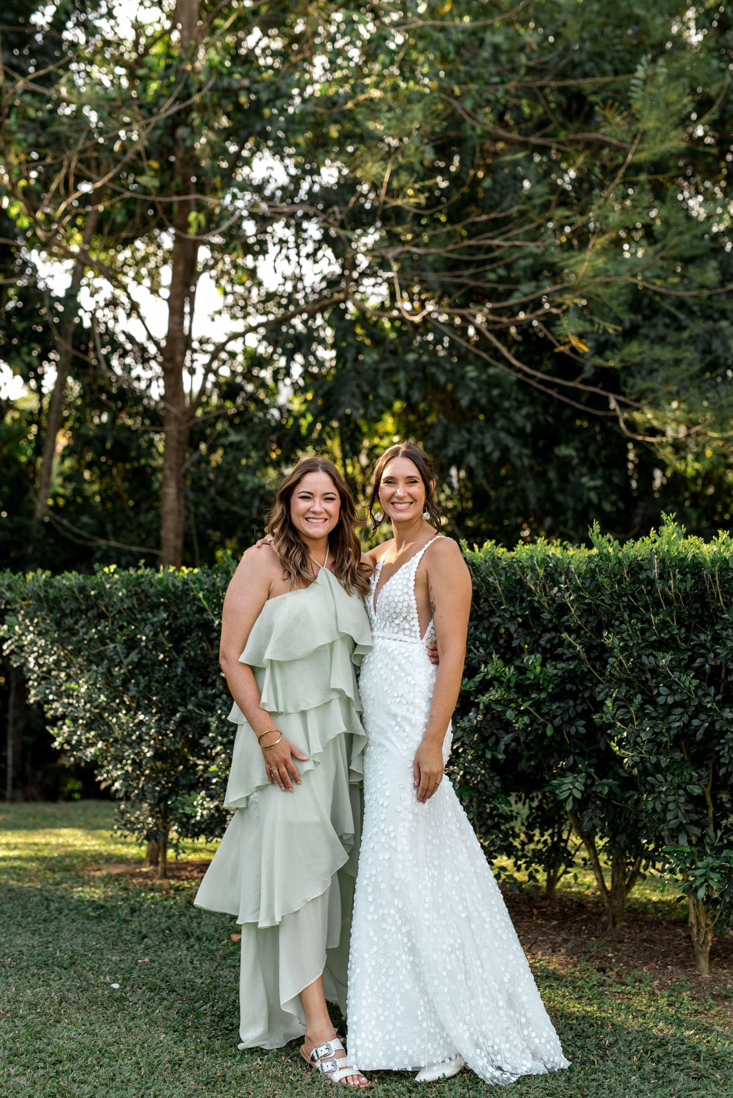 Bride in white lace gown poses with bridesmaid in sage green ruffle dress at outdoor garden wedding.