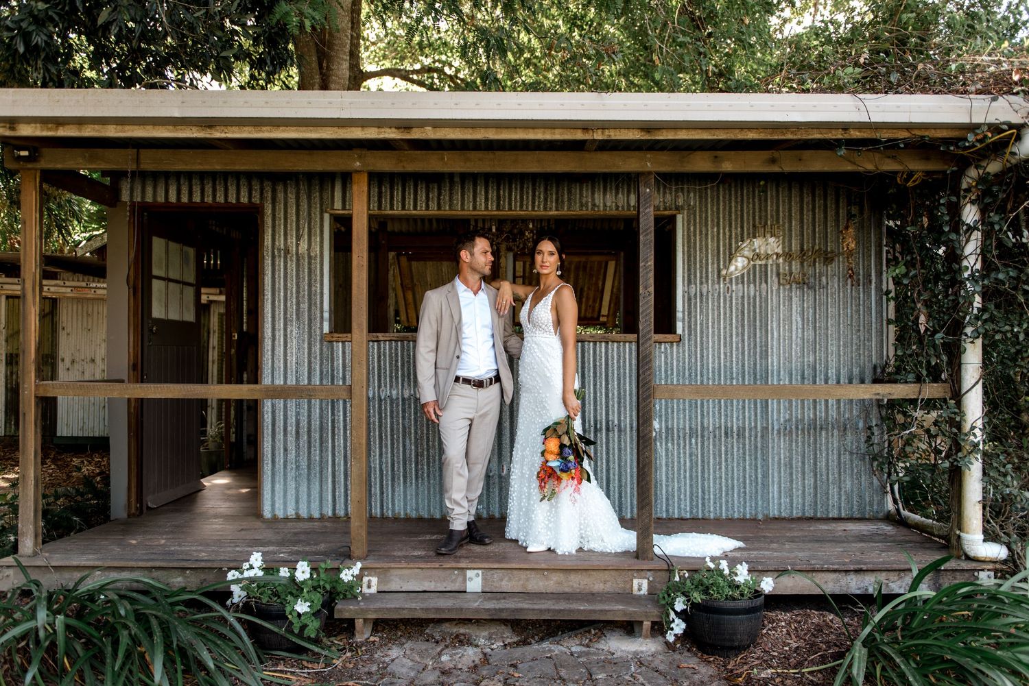 Bride and groom pose on a charming rustic corrugated iron cottage porch with bouquet.