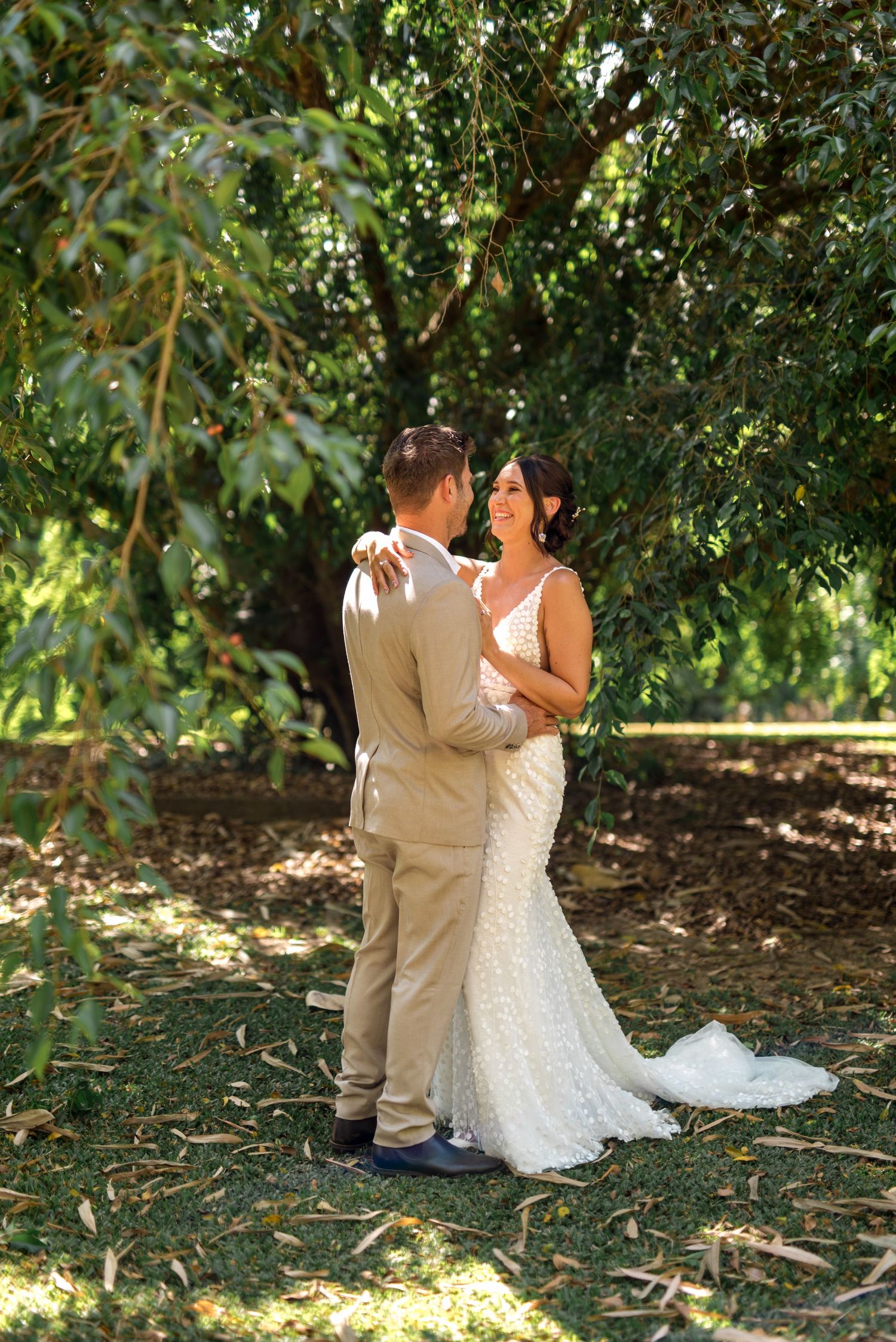 Wedding couple share a romantic moment beneath a canopy of green trees in natural light.