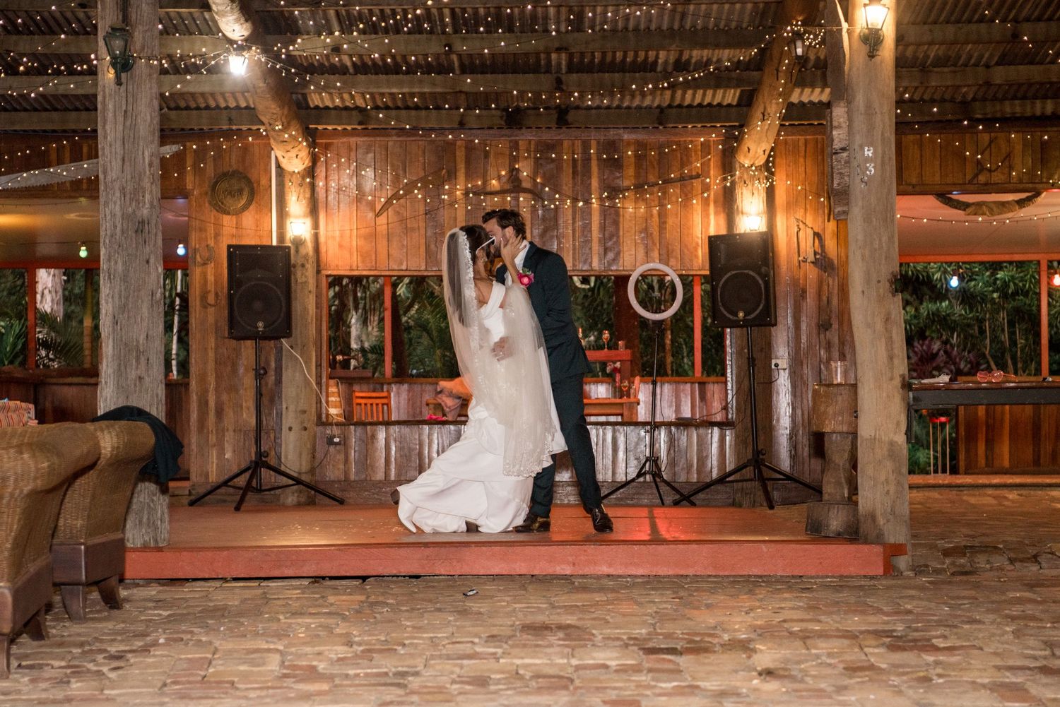 Bride and groom share their first dance on a rustic wooden stage adorned with warm string lights.