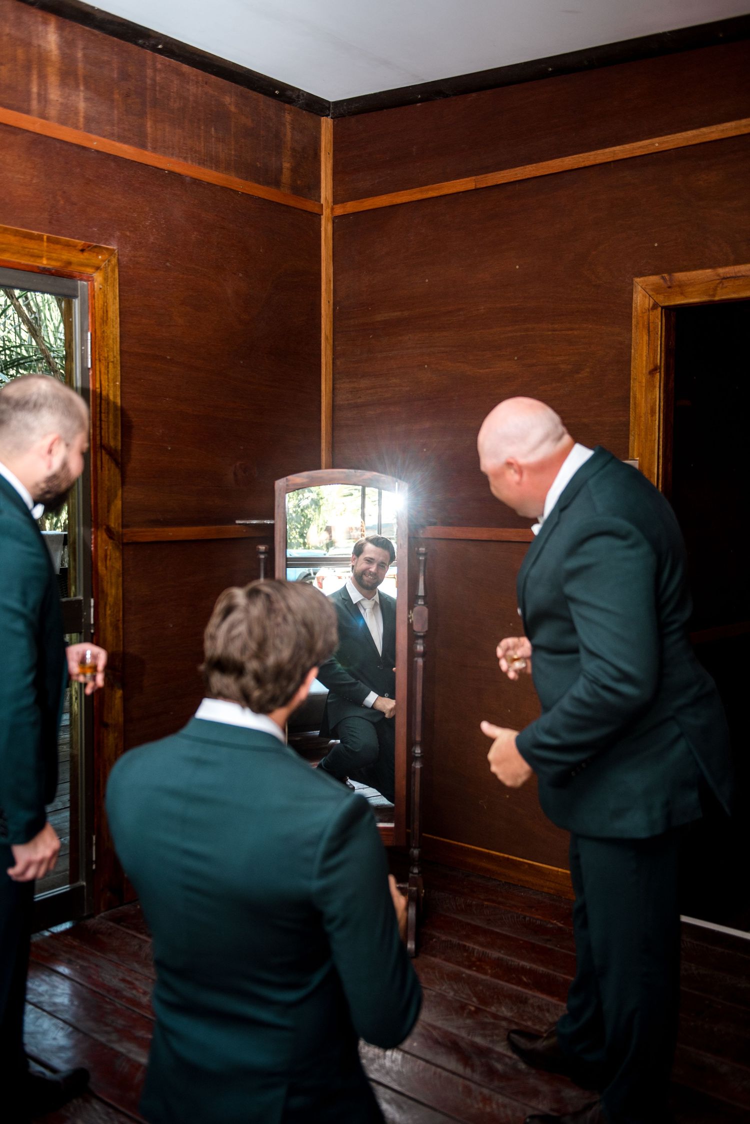 Groomsmen helping each other get ready in rustic wooden room with mirror.