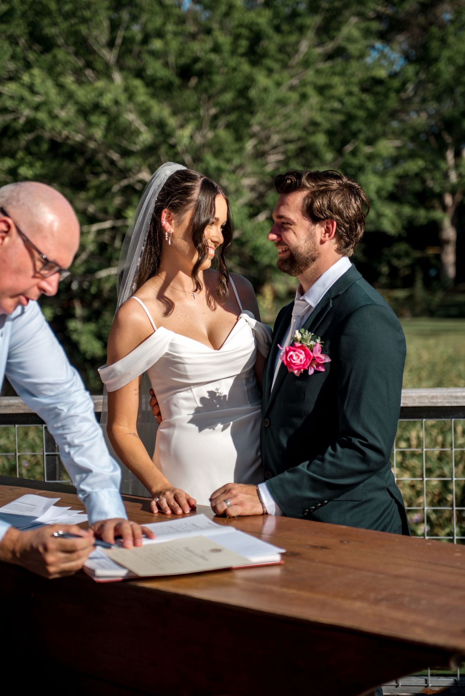 Bride and groom smiling while signing marriage documents outdoors.