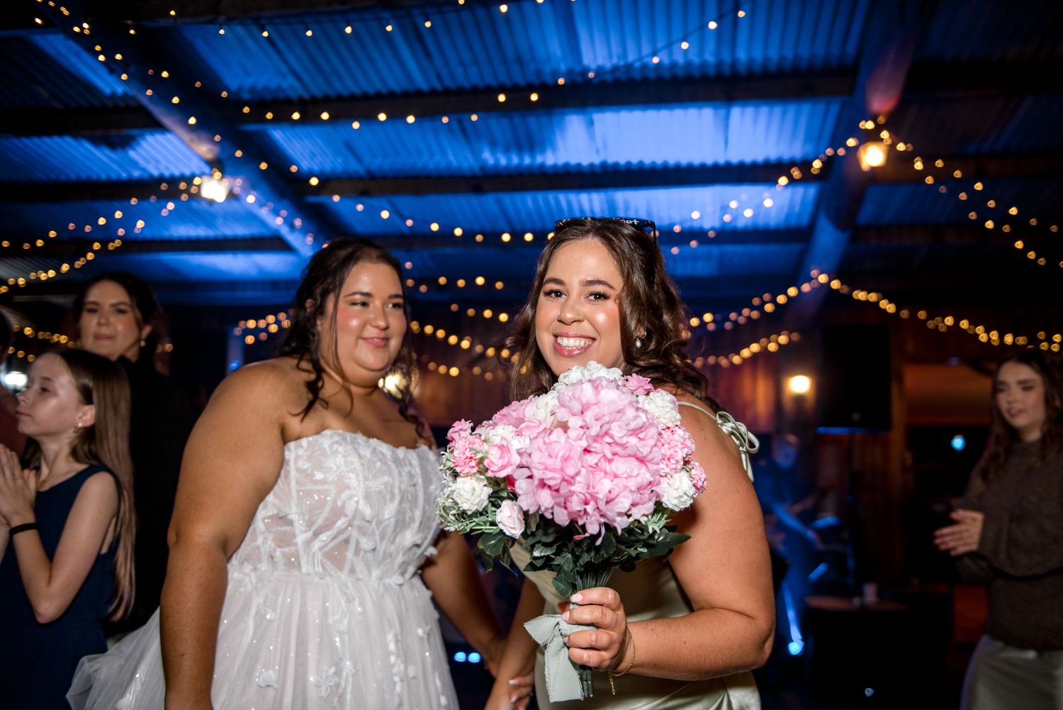 Smiling bride holding pink roses bouquet with guest at wedding reception with blue lighting.