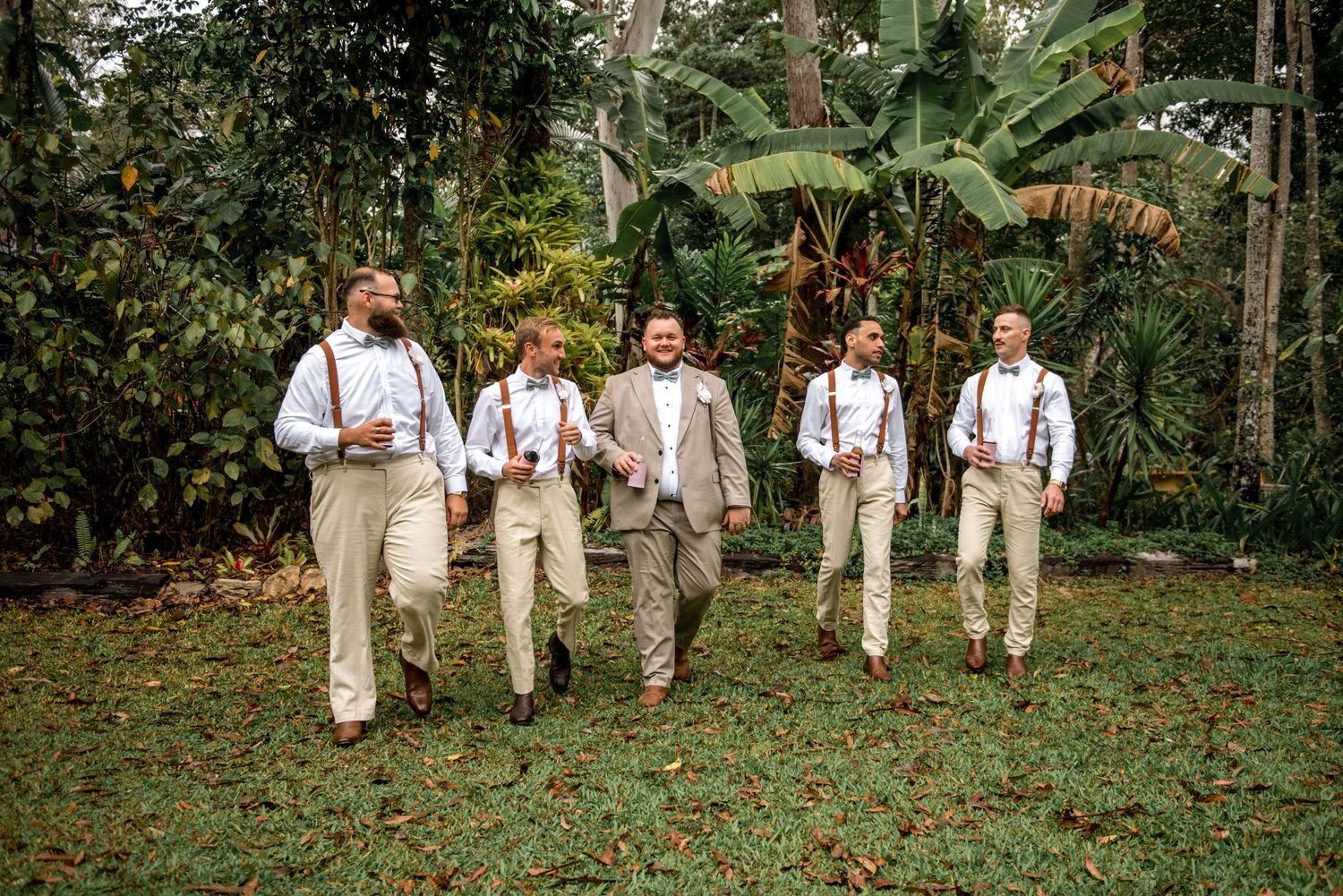 Groom in tan suit leads groomsmen in white shirts and suspenders walking through a tropical garden.