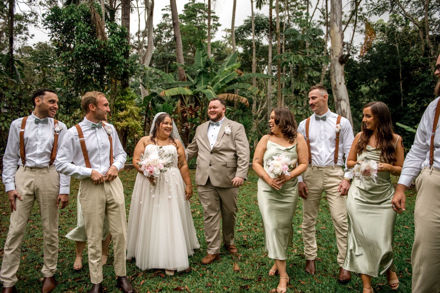 Bride and groom walk with wedding party in sage green dresses and tan suits in lush garden.