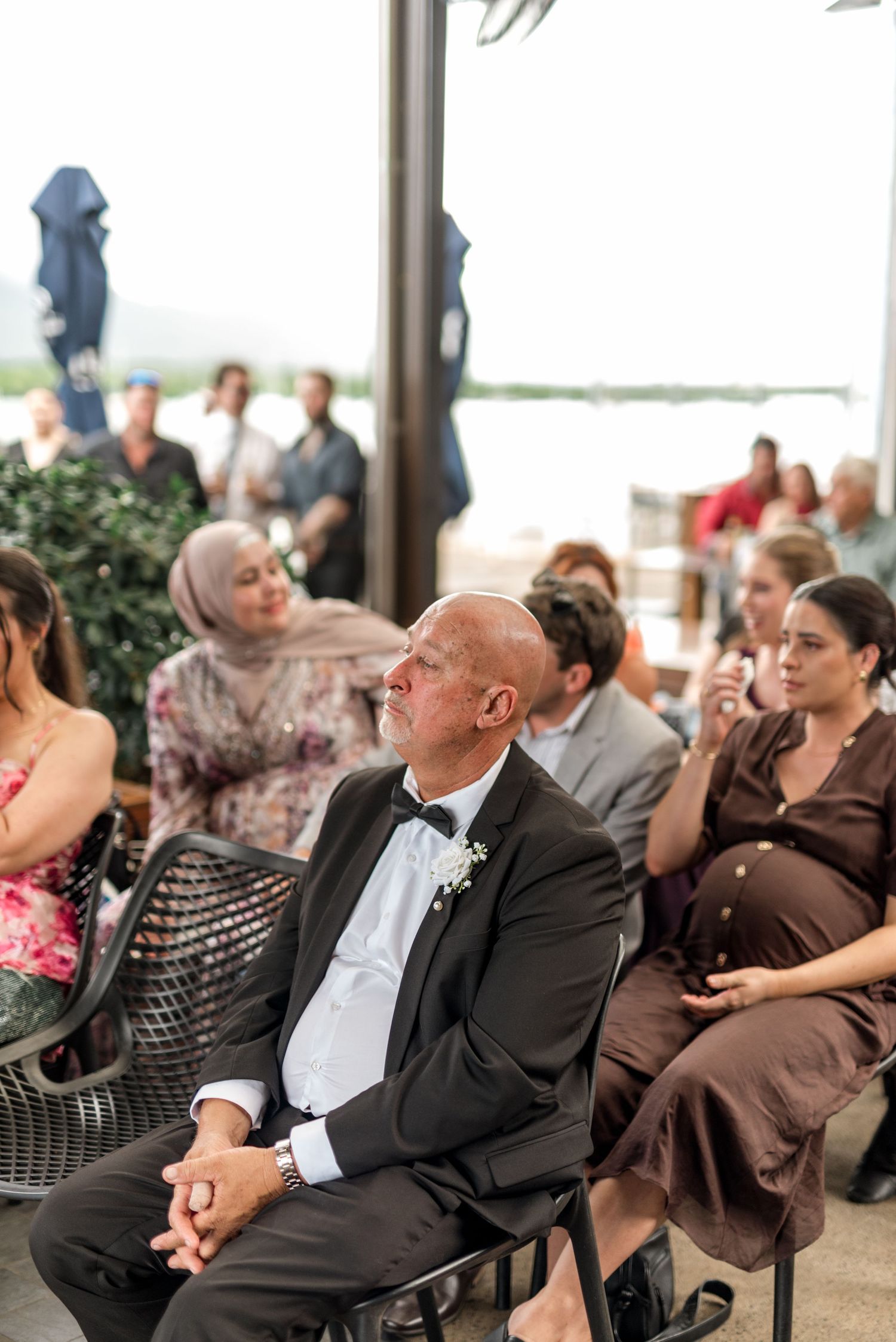 Emotional wedding guest in bow tie watches ceremony with other guests at waterfront venue.
