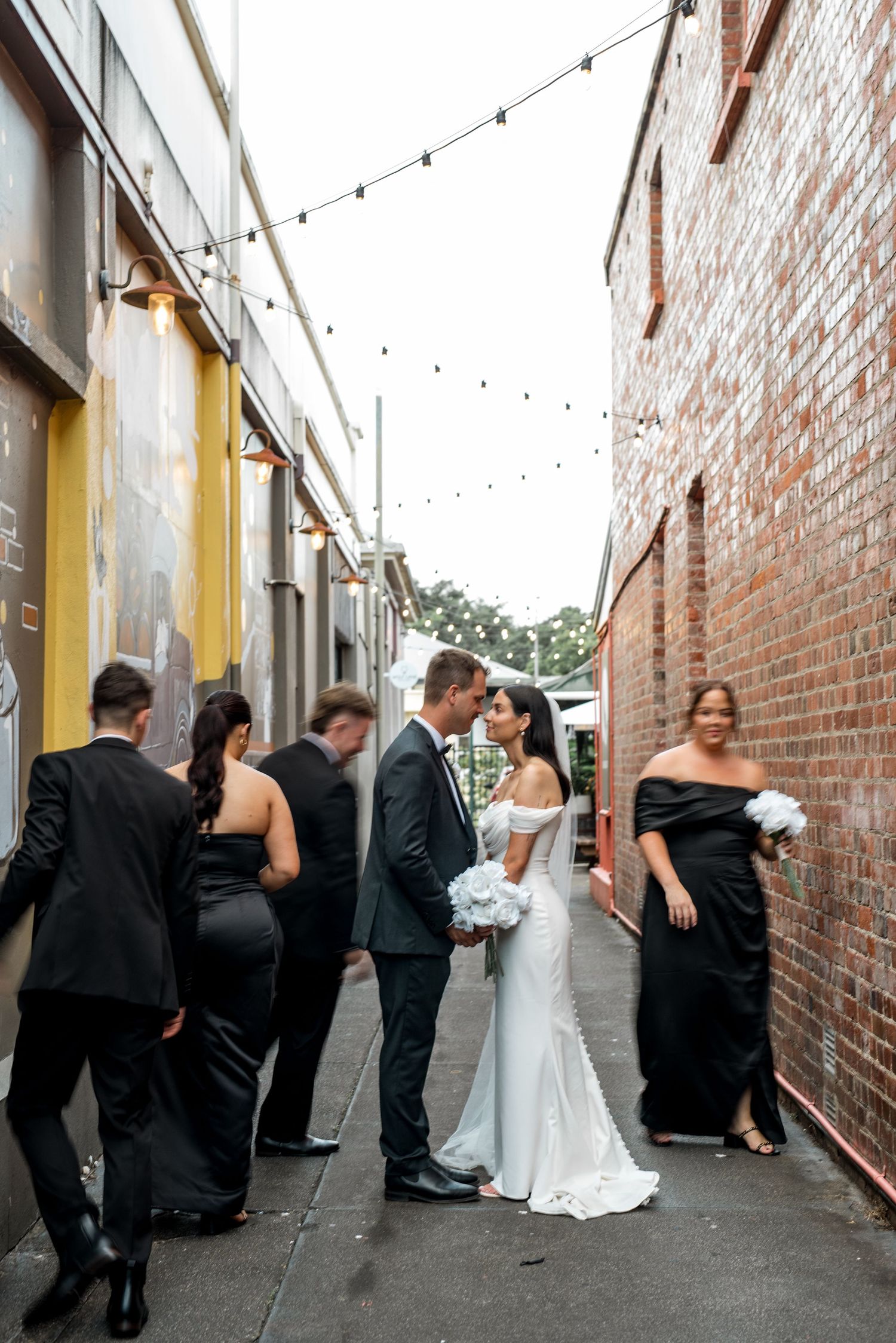 Couple kissing in a brick alleyway as wedding party mingles around them.