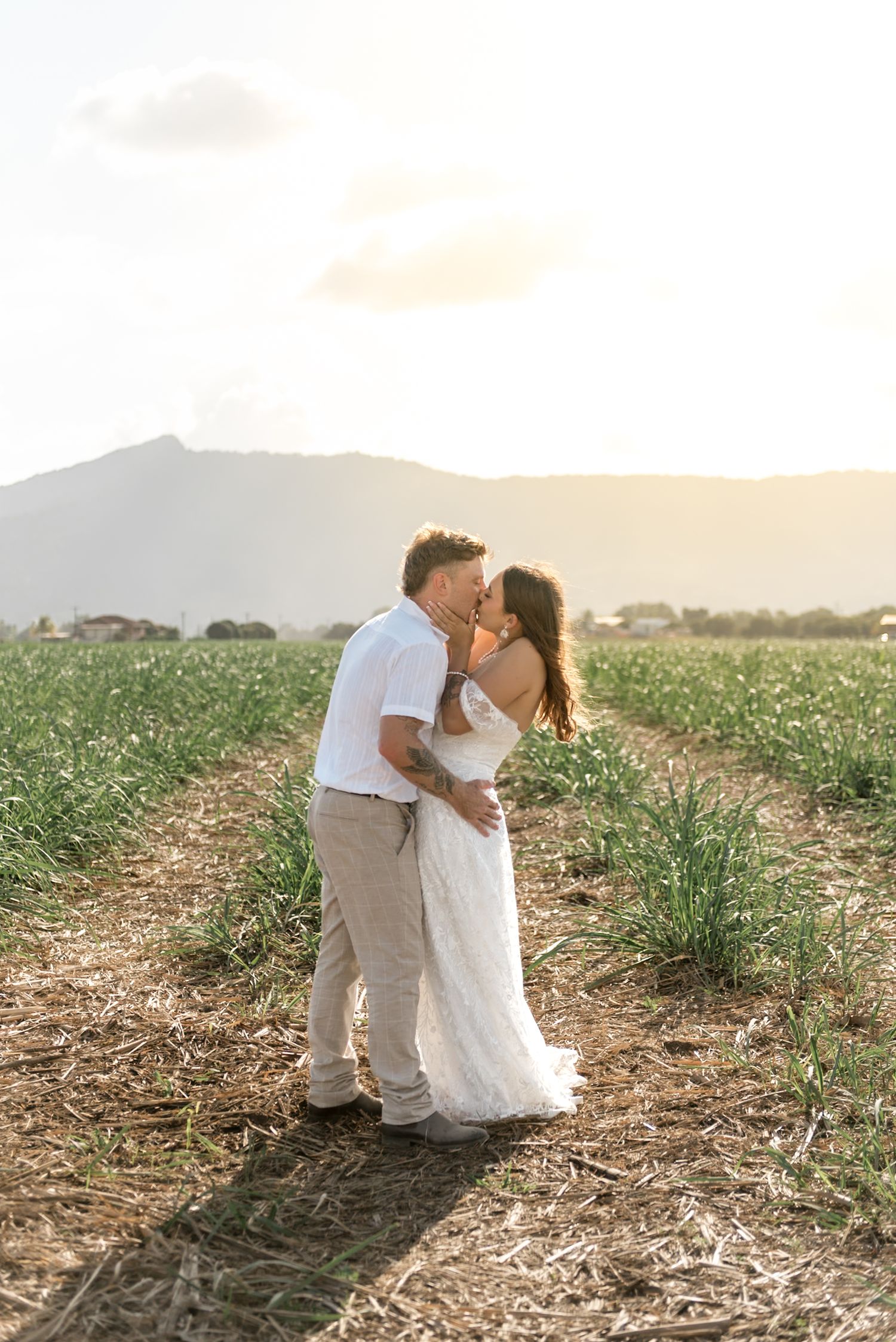 Newlyweds kissing at golden hour in a green field with stunning mountain views in background.