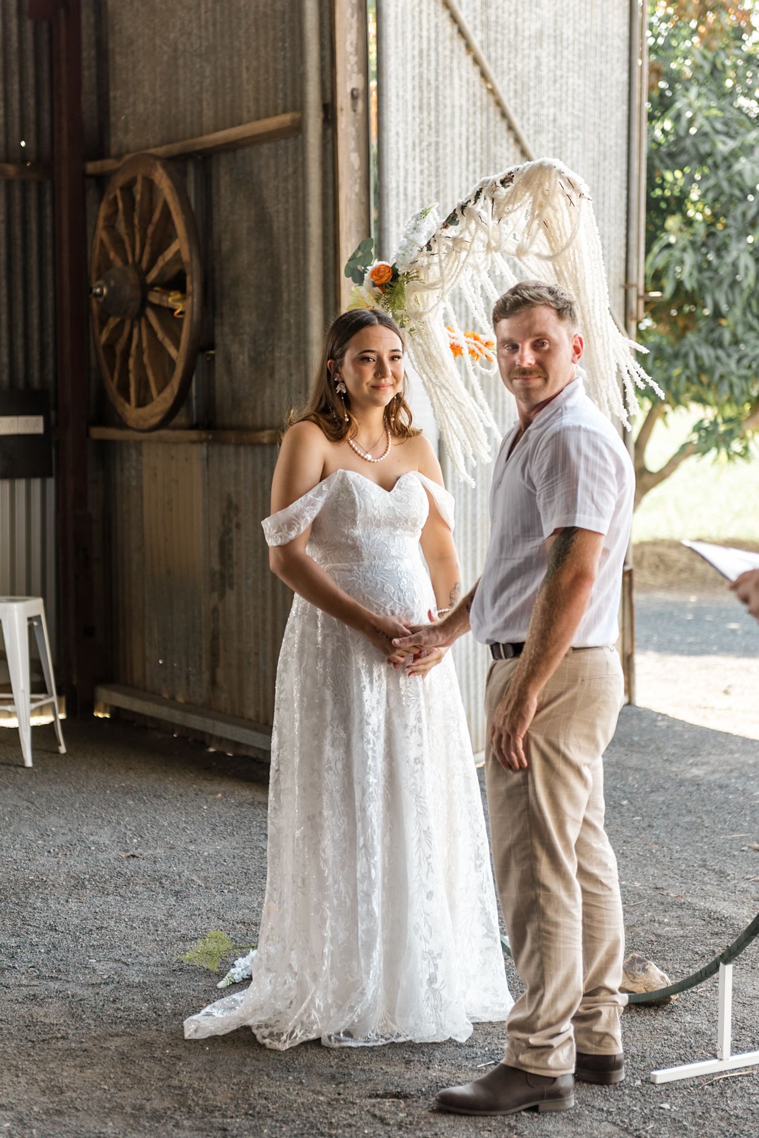 Bride and groom pose together under boho macrame arch in rustic barn wedding venue with wagon wheel decor.