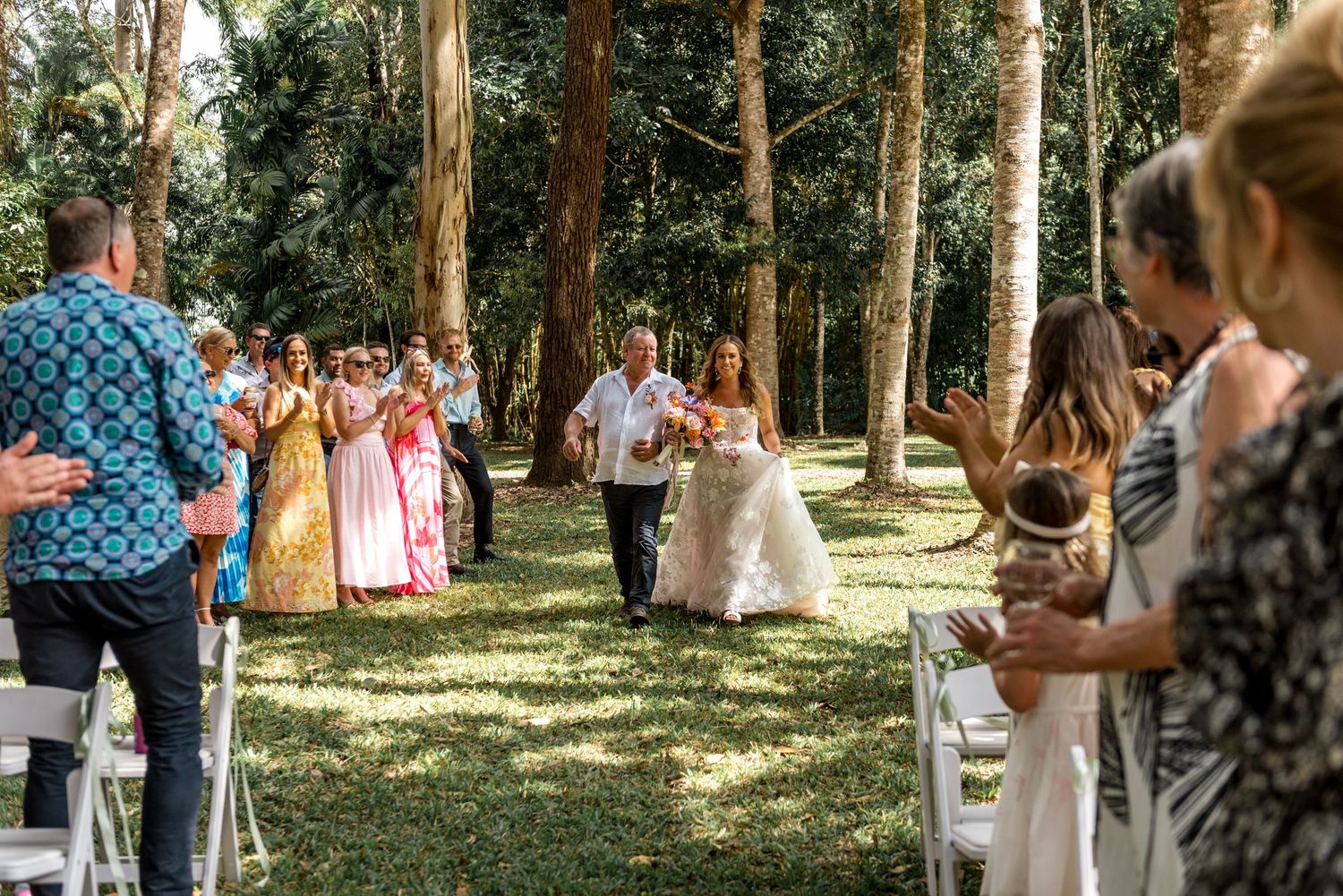 Bride walking down the aisle with her escort in a sunlit forest wedding ceremony.