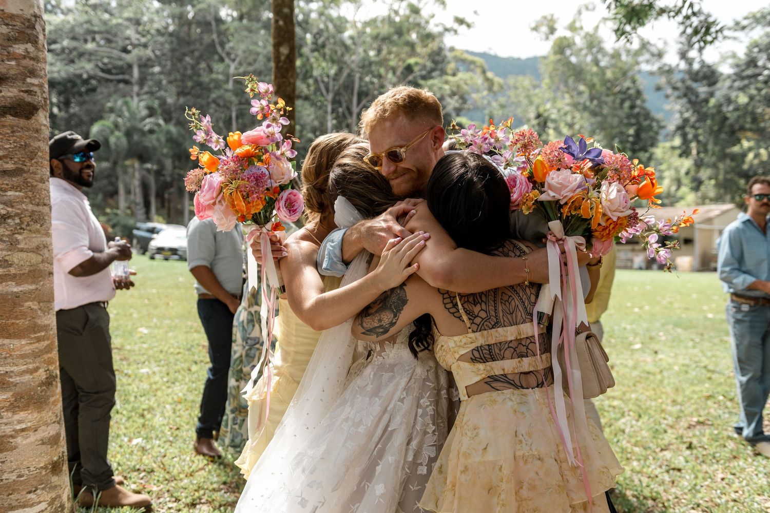 Bride and groom sharing a group hug with bridesmaids holding colorful bouquets at outdoor wedding.