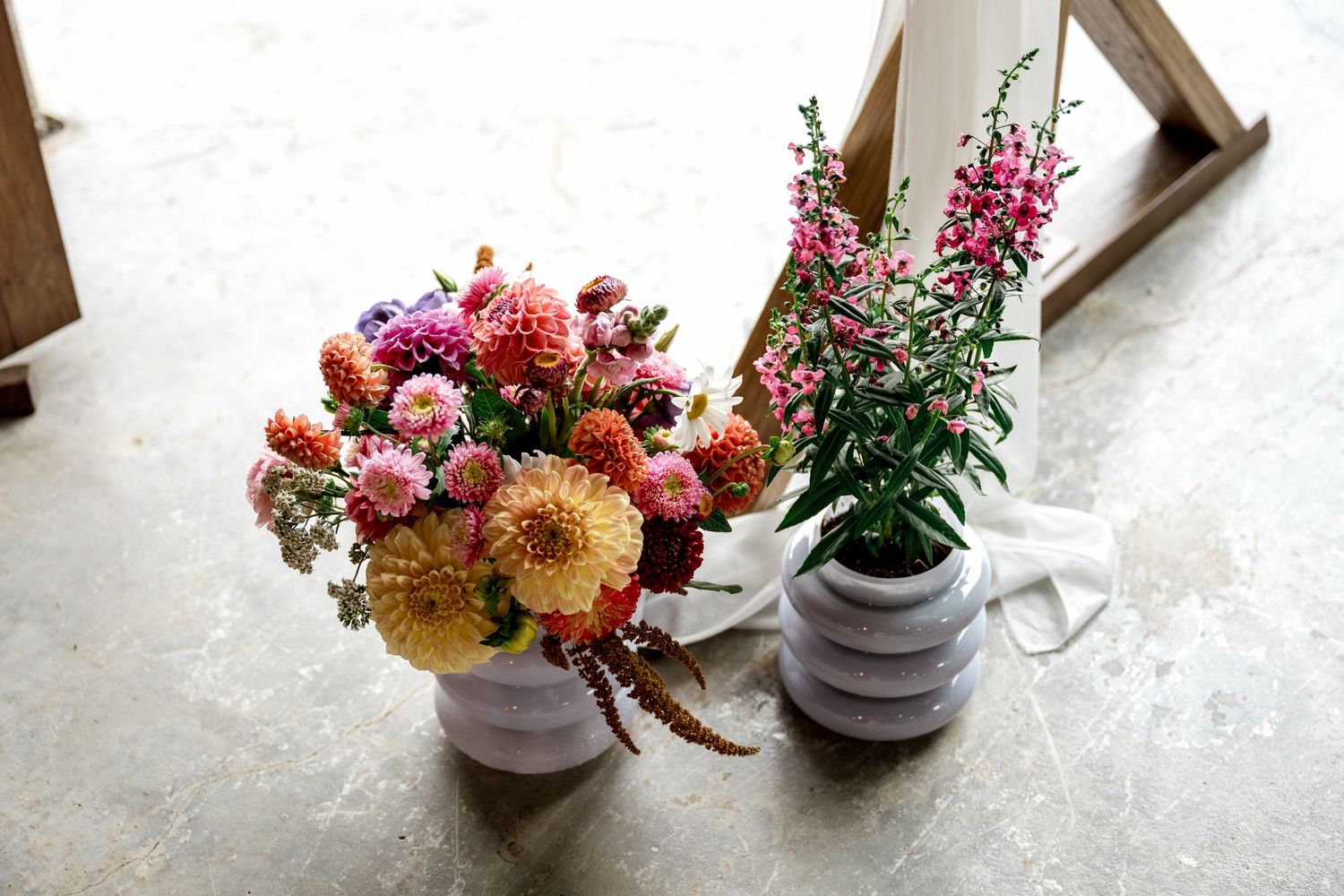 Two floral arrangements with pink dahlias and tall magenta flowers in white vases on concrete floor.