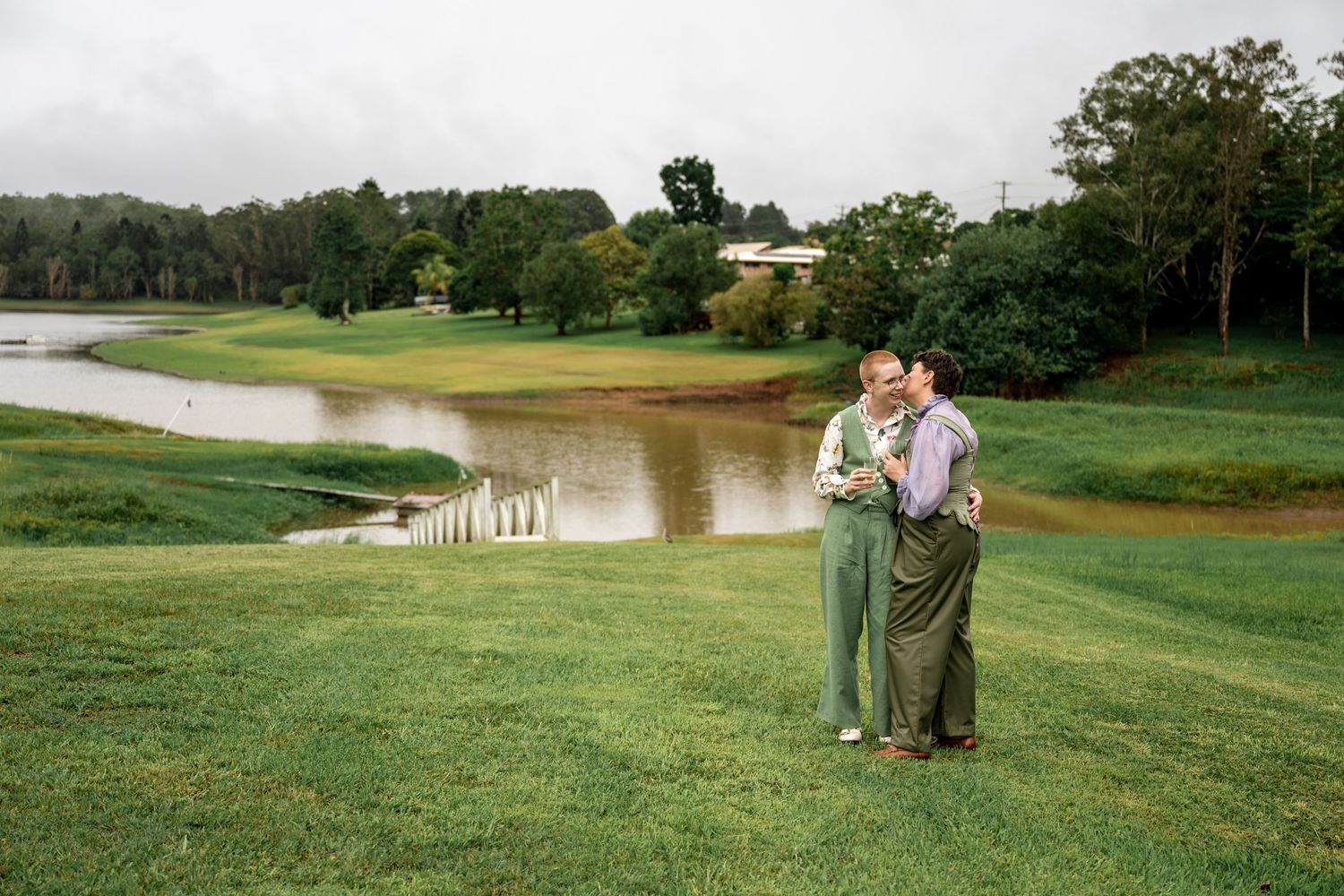 Couple sharing a romantic kiss on lush green lawn with tranquil pond backdrop.