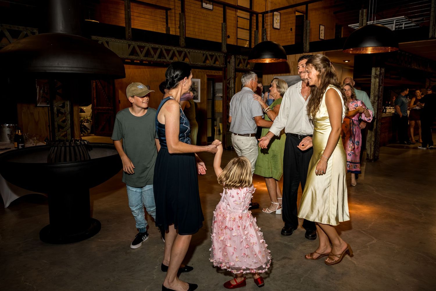 Wedding guests including a young girl in pink dance together at a rustic venue.