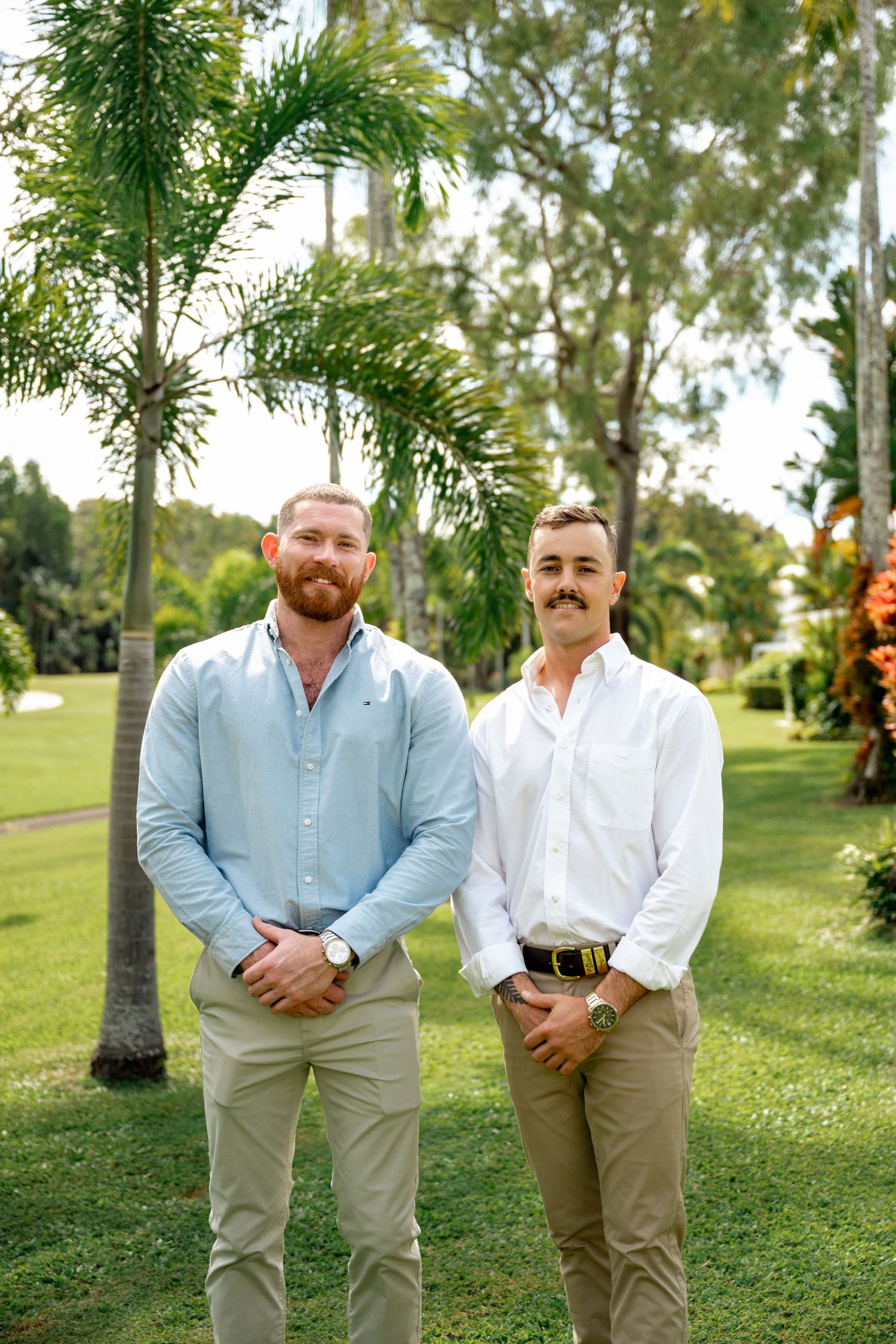 Two men in light blue and white shirts posing together on green lawn with tropical palms.