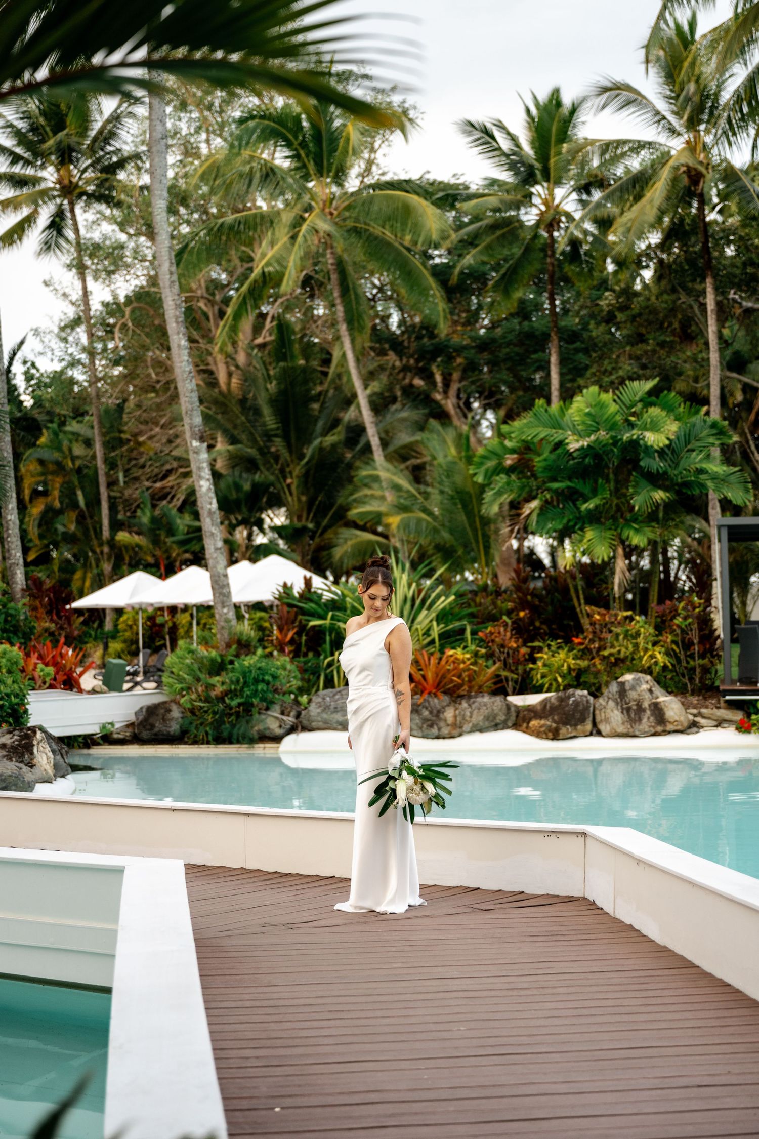 Bride in a sleek white gown standing on a wooden deck by a tropical resort pool with palm trees at Sheraton Grand Mirage Port Douglas.