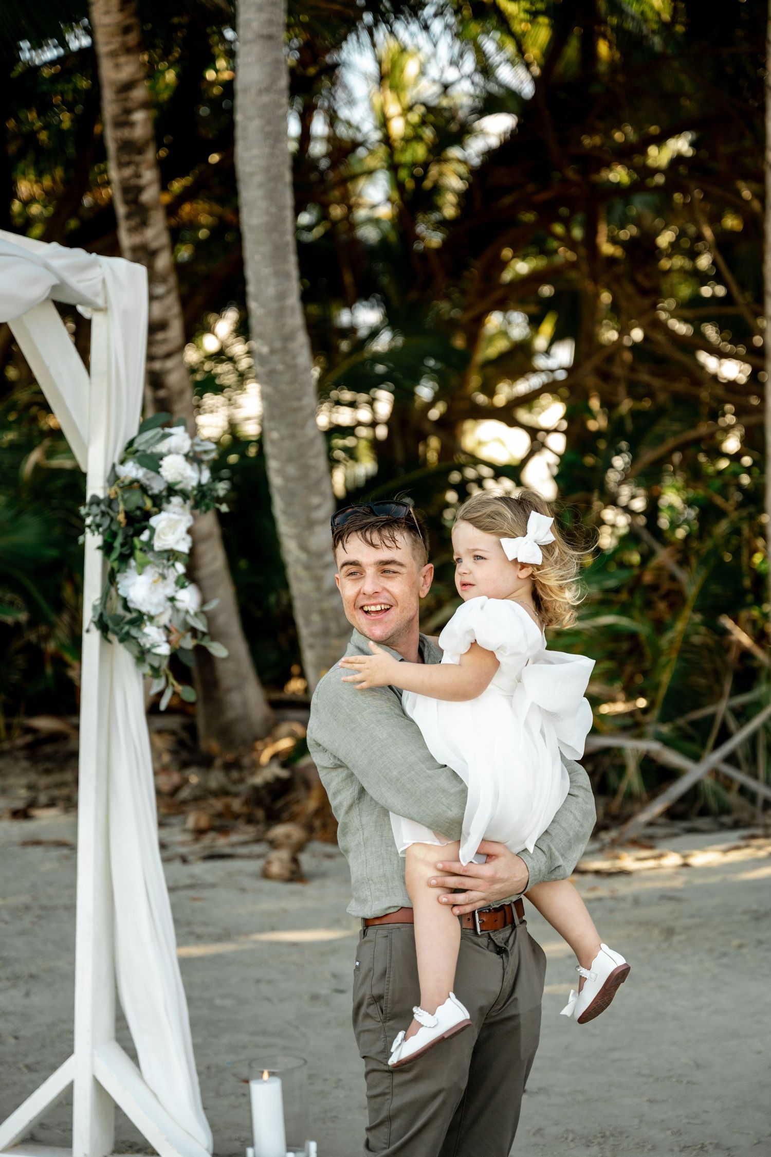 Man in gray shirt holds smiling toddler girl in white dress at a beach wedding ceremony with floral arch at Four Mile Beach, Port Douglas.
