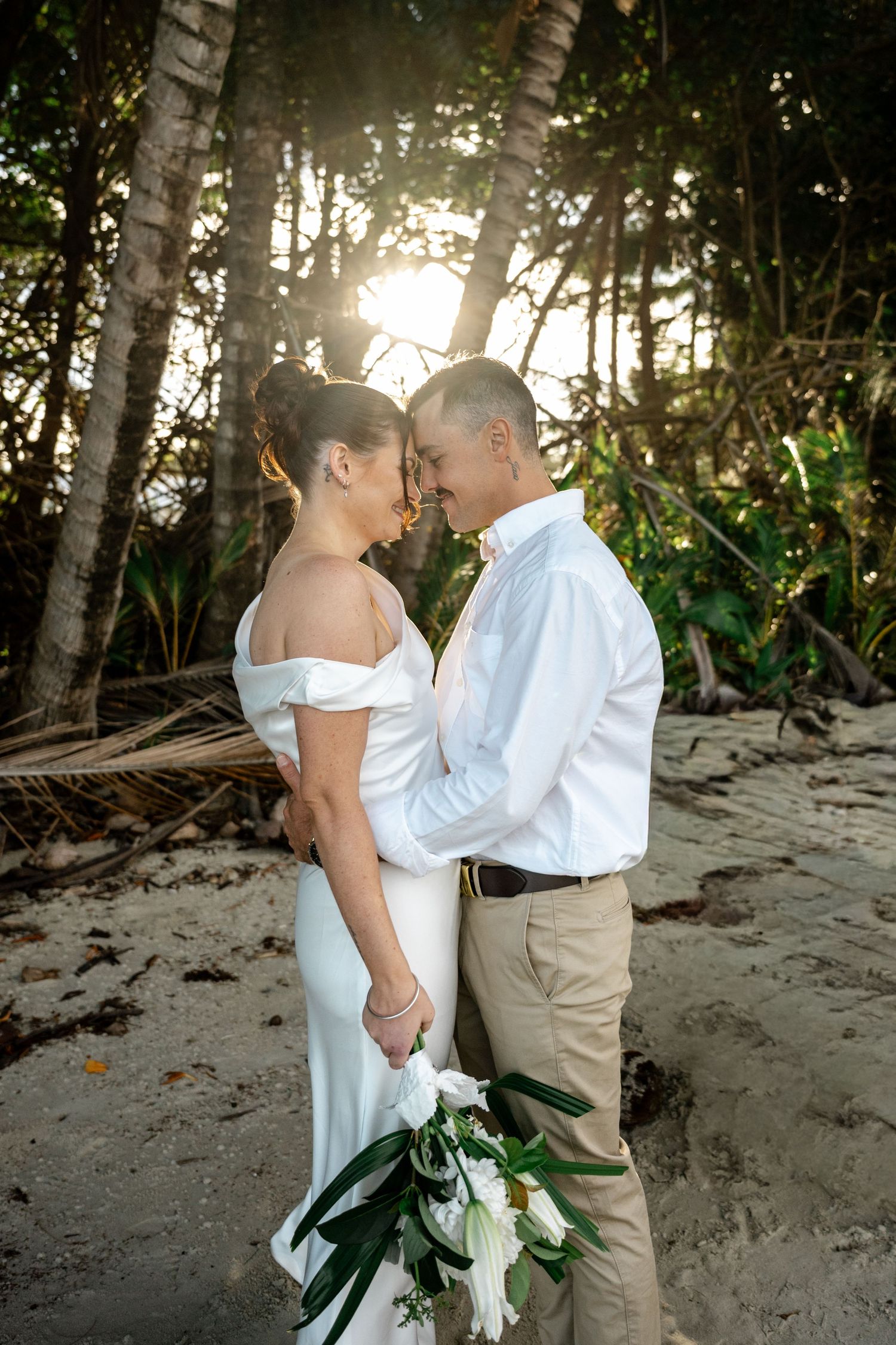 Newlyweds share an intimate moment holding a white bouquet surrounded by palm trees at sunset at Four Mile Beach, Port Douglas.