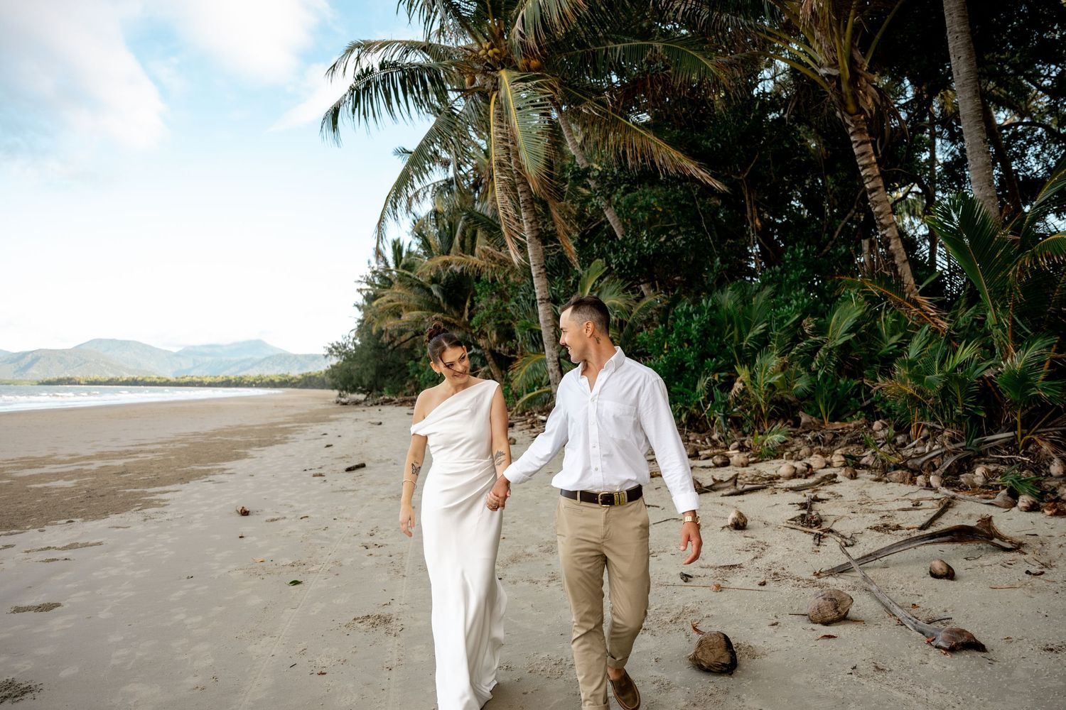 Smiling bride and groom walk hand in hand along a tropical beach with palm trees at Four Mile Beach, Port Douglas.
