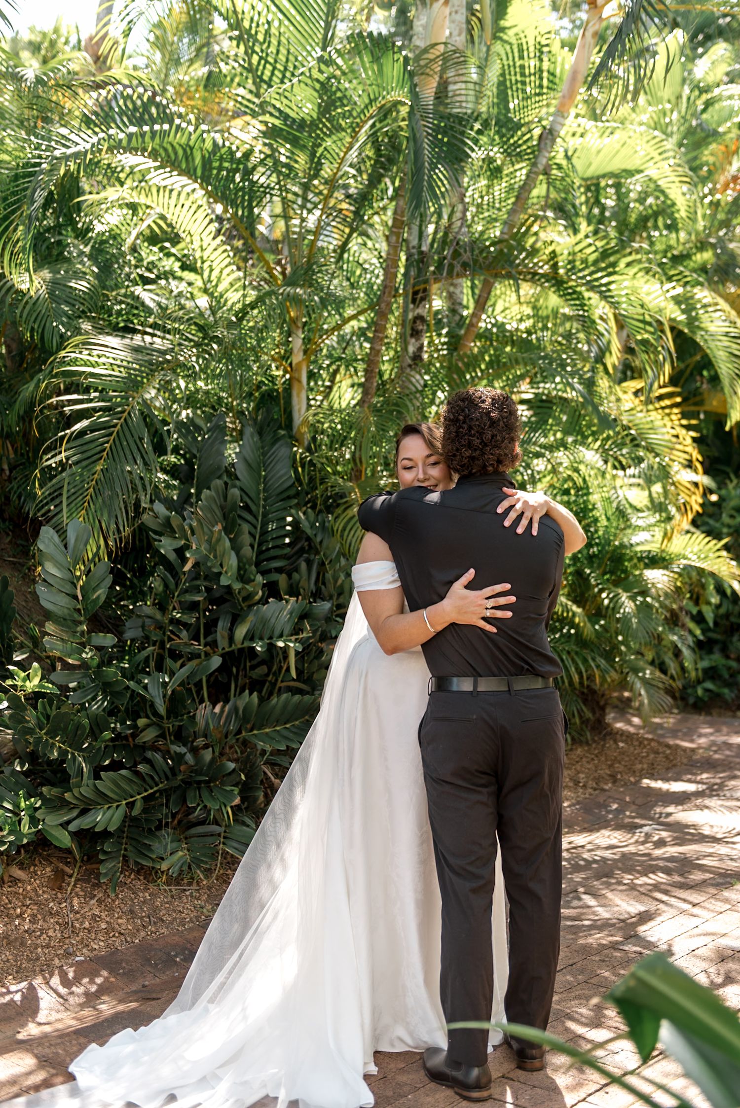 Bride and groom share tender embrace surrounded by lush tropical palm trees on wedding day.