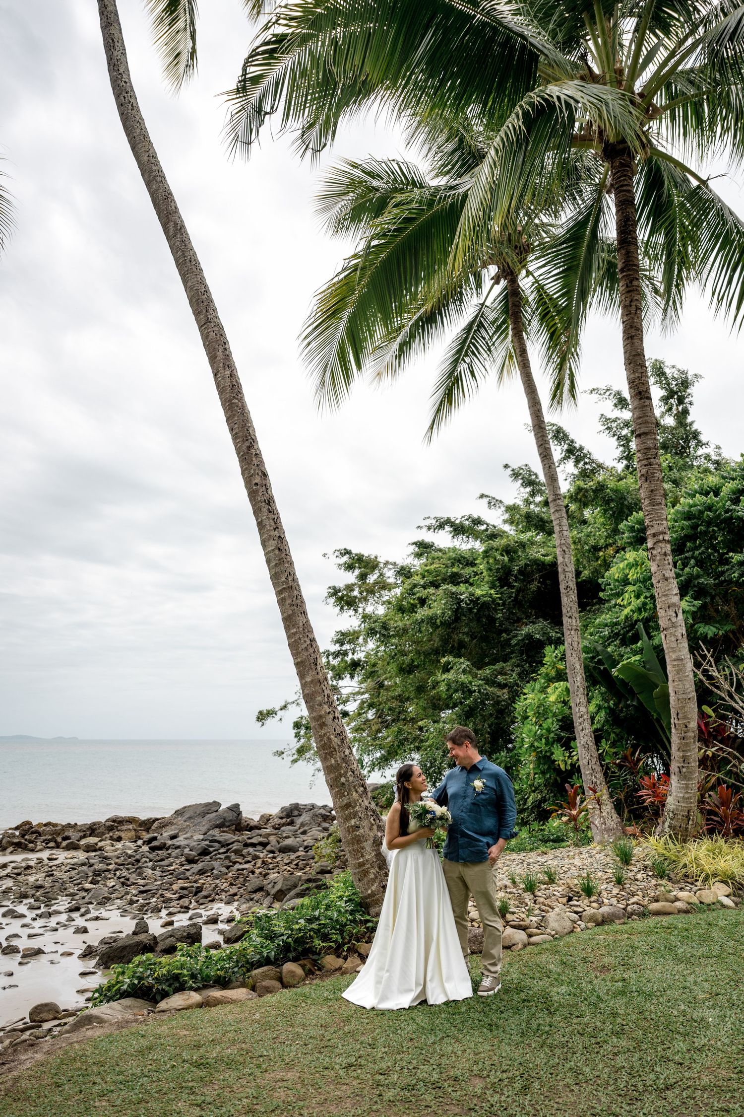 Bride and groom stand romantically beneath tall palm trees on a tropical beachside lawn on their wedding day at Little Cove, Port Douglas.