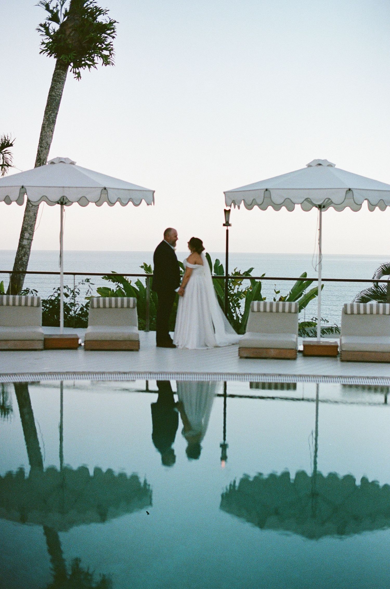 Bride and groom reflected in a calm pool at twilight with white beach umbrellas.